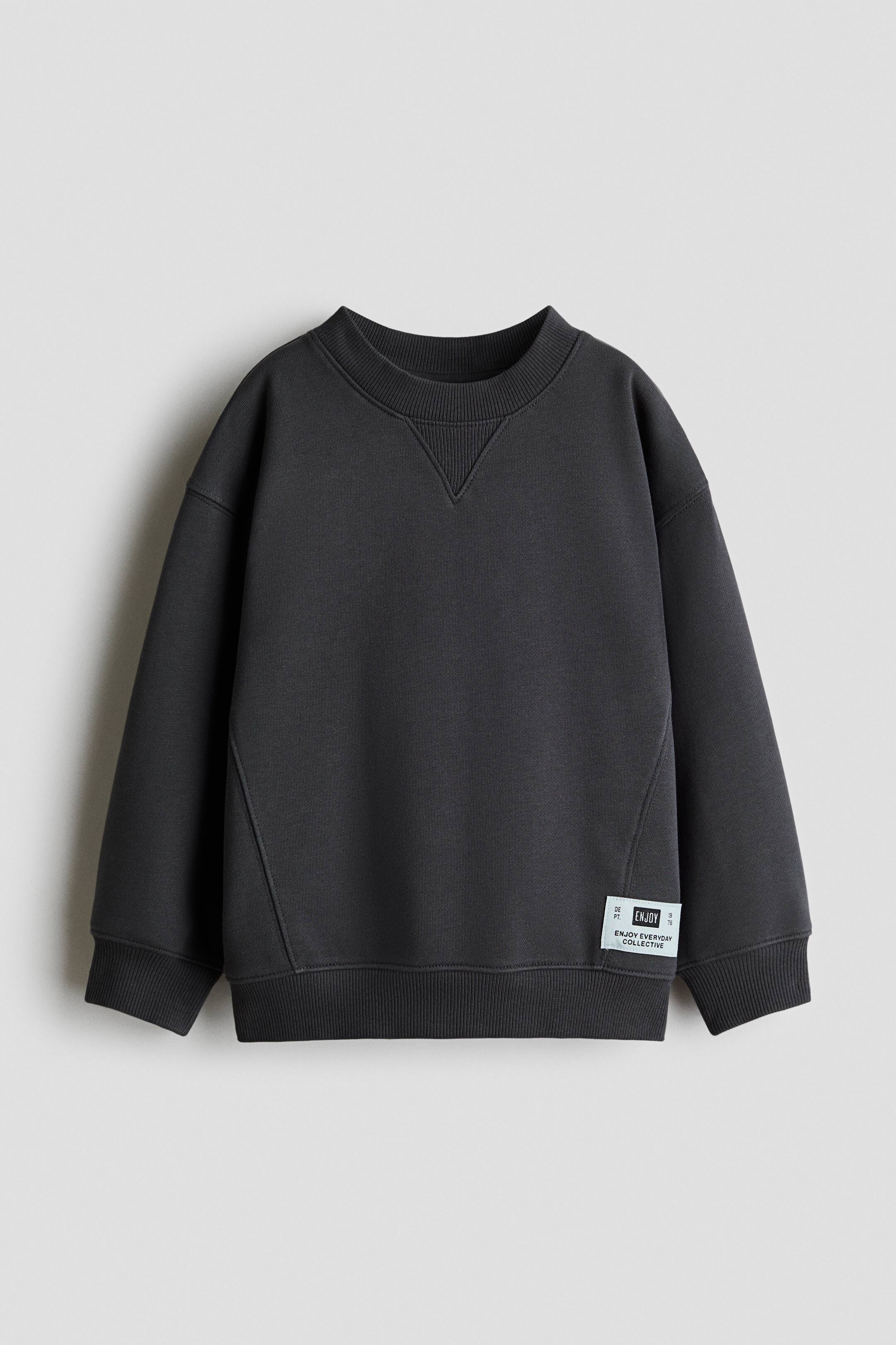 Ampliar la imagen: Sudadera de algodón gris oscuro vista de frente, con cuello redondo acanalado y detalle de costura en forma de V, mangas largas con puños acanalados y dobladillo acanalado. Las costuras anguladas son visibles en la parte frontal del torso, y una pequeña etiqueta tejida está adherida al lado izquierdo cerca del dobladillo.