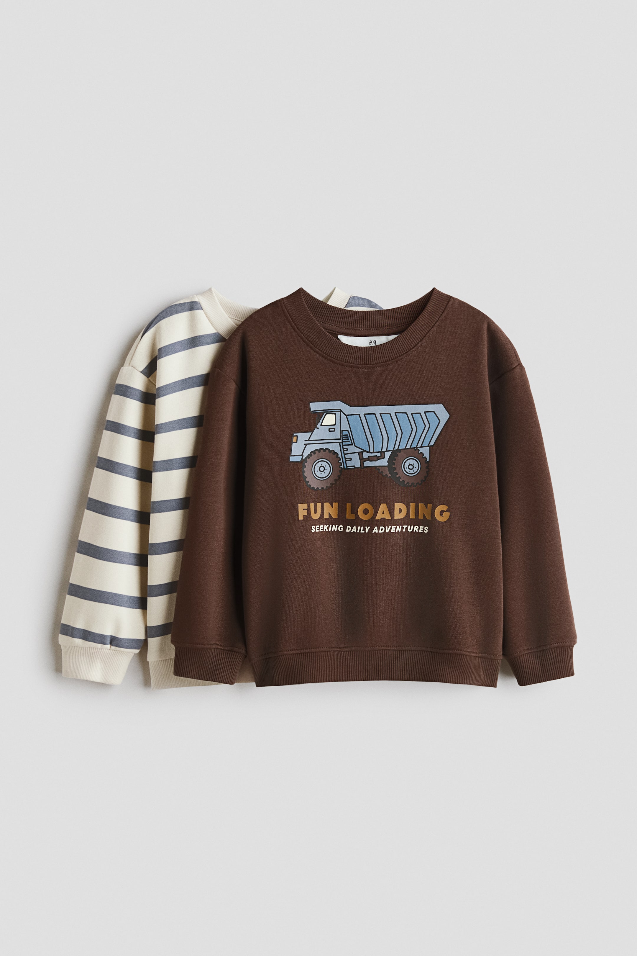 View larger image: Long-sleeved tops in printed fleece sweatshirt fabric, with the front-facing brown sweatshirt displaying a dump truck graphic and 'fun loading seeking daily adventures' text, and a partially visible cream sweatshirt with blue-grey horizontal stripes positioned behind it. Both tops feature round, rib-trimmed necklines, dropped shoulders, long sleeves, and ribbing at the cuffs and hem.