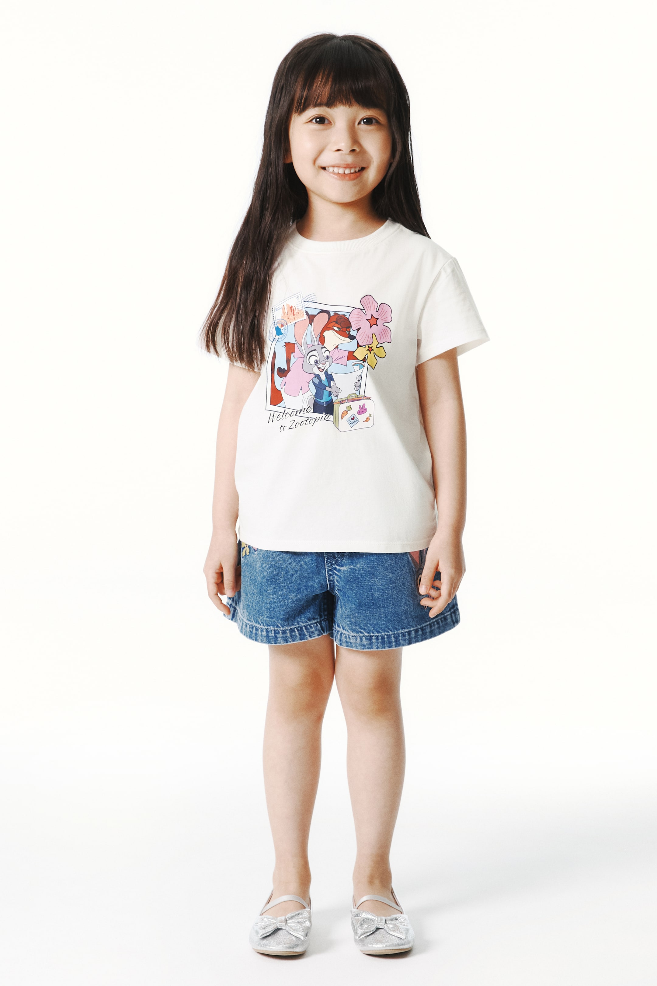 이미지 확대 보기: 모델이 착용한 티셔츠는 부드러운 프린트 면 저지 소재로, 둥근 리브 트림 넥라인이 특징입니다. 티셔츠 앞면에는 애니메이션 캐릭터와 꽃이 어우러진 컬러풀한 그래픽이 프린트되어 있습니다.