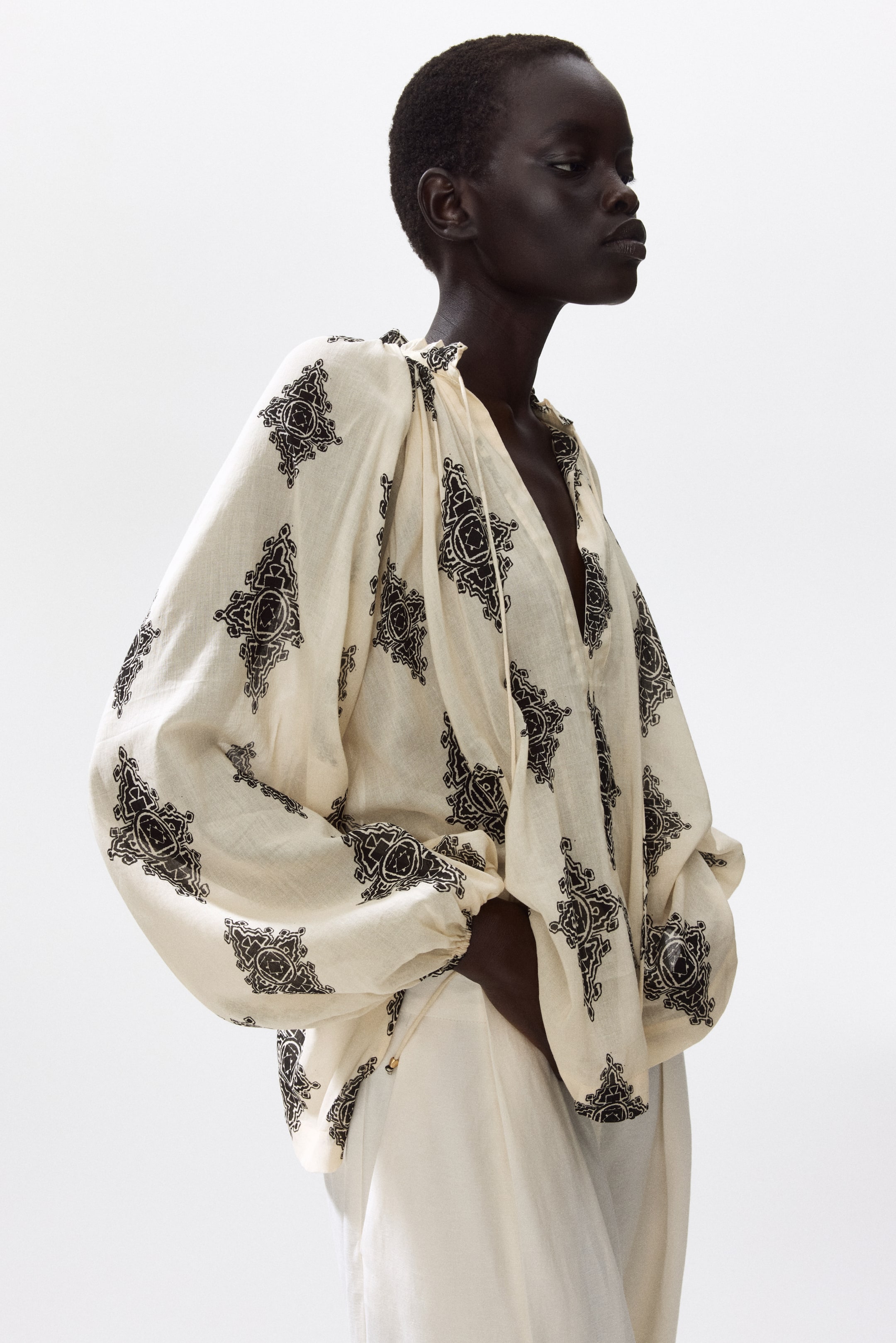 Agrandir l'image: Un chemisier crème oversize avec un imprimé noir est porté par le mannequin, doté d'un col à volants froncés avec une fente en V profond, de liens spaghetti, de longues manches ballon volumineuses en coupe raglan et de poignets élastiqués. Le mannequin porte également un pantalon de couleur claire, avec sa main posée sur sa hanche.