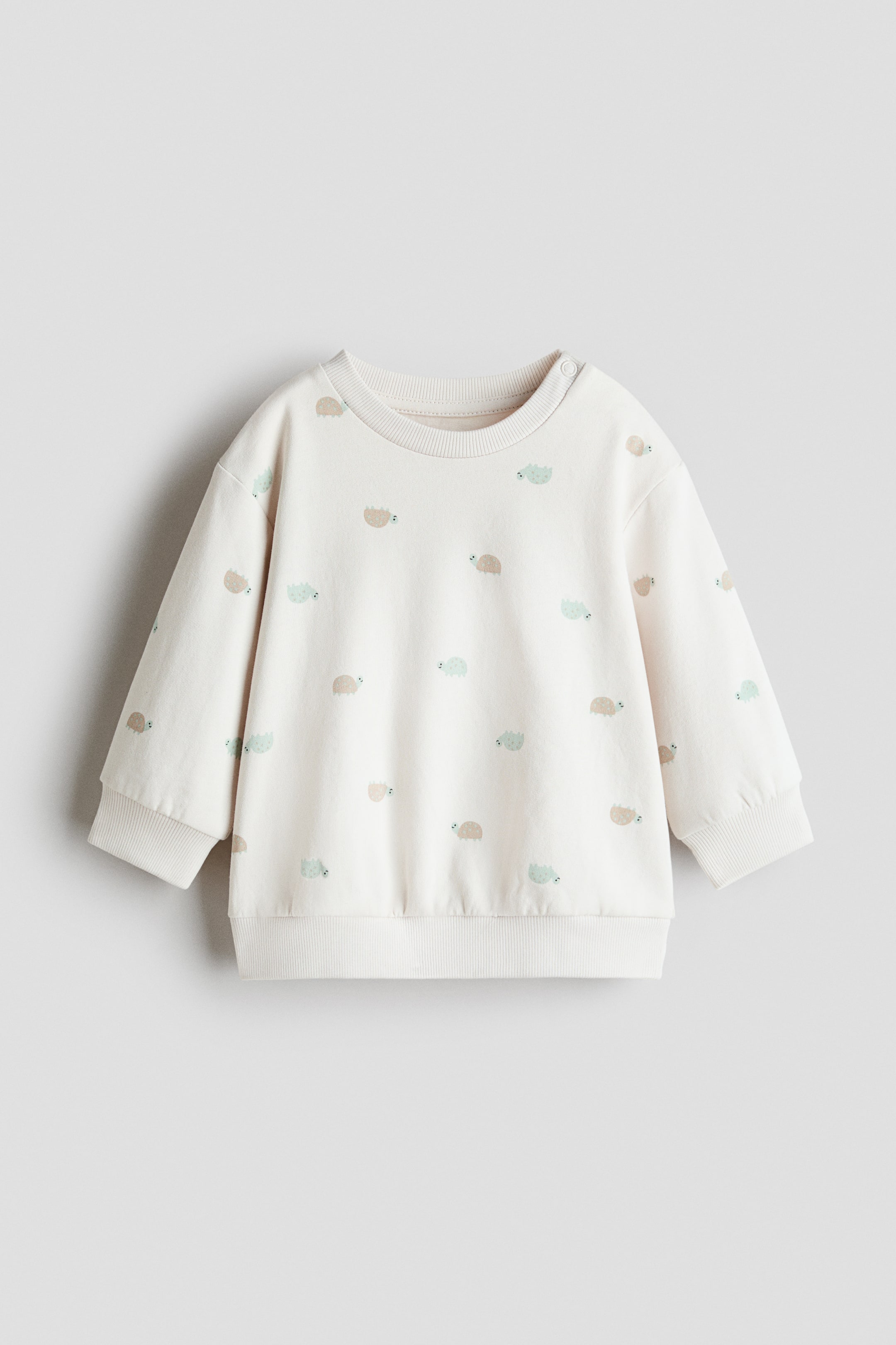 Büyük resmi görüntüle: Krem renkli bir sweatshirt önden gösterilmektedir; küçük, stilize açık kahverengi ve açık yeşil hayvan motiflerinin dağınık desenine sahiptir. Yuvarlak, kanal örgülü bir yaka, sağ omuzda tek bir çıtçıt, kanal örgülü manşetler ve kanal örgülü etek ucu ile tasarlanmıştır.
