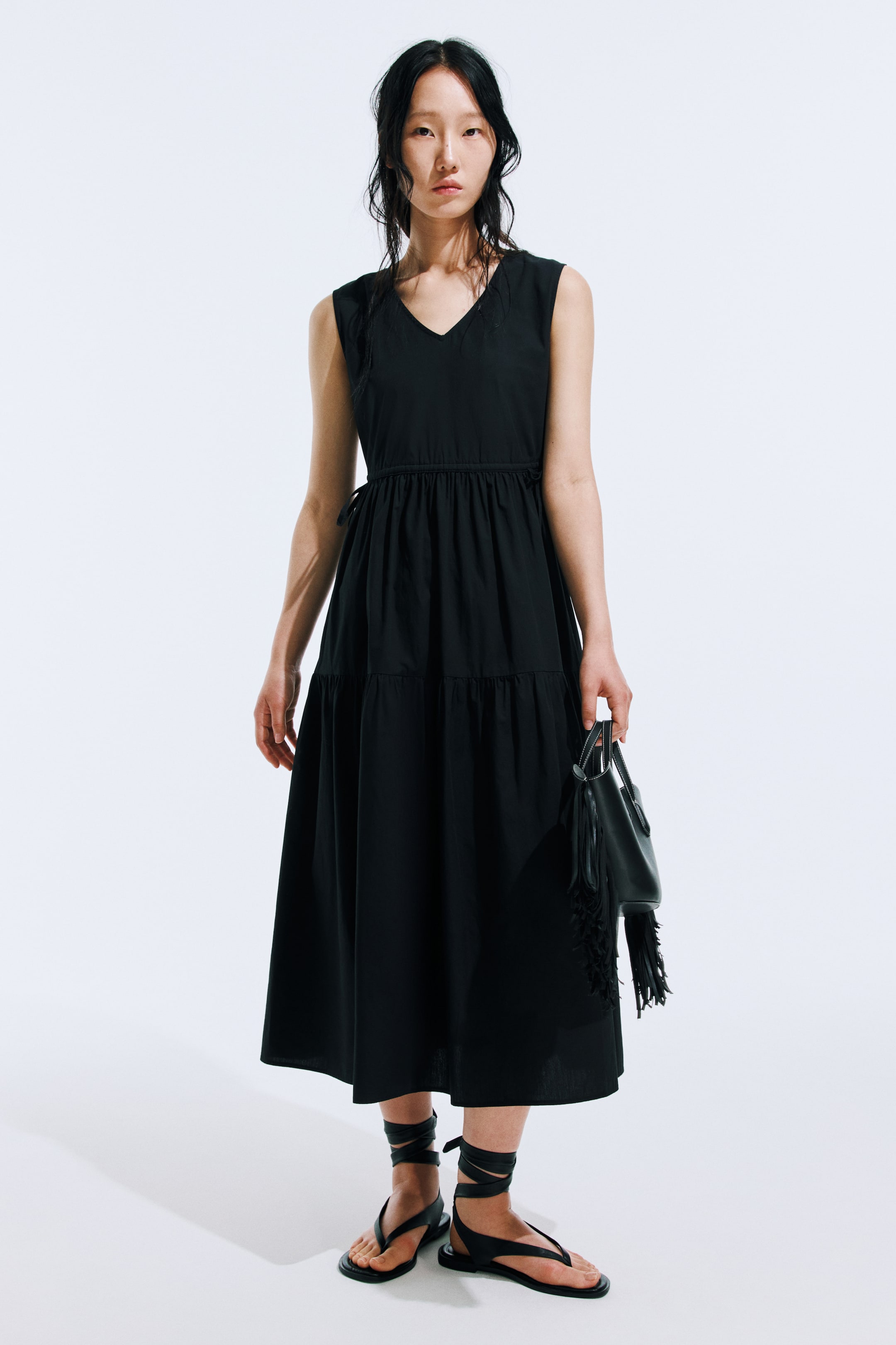 View larger image: The model is wearing a sleeveless black dress that falls to calf-length, showcasing a V-neckline, a drawstring waist, and a flared skirt with a gathered tier.