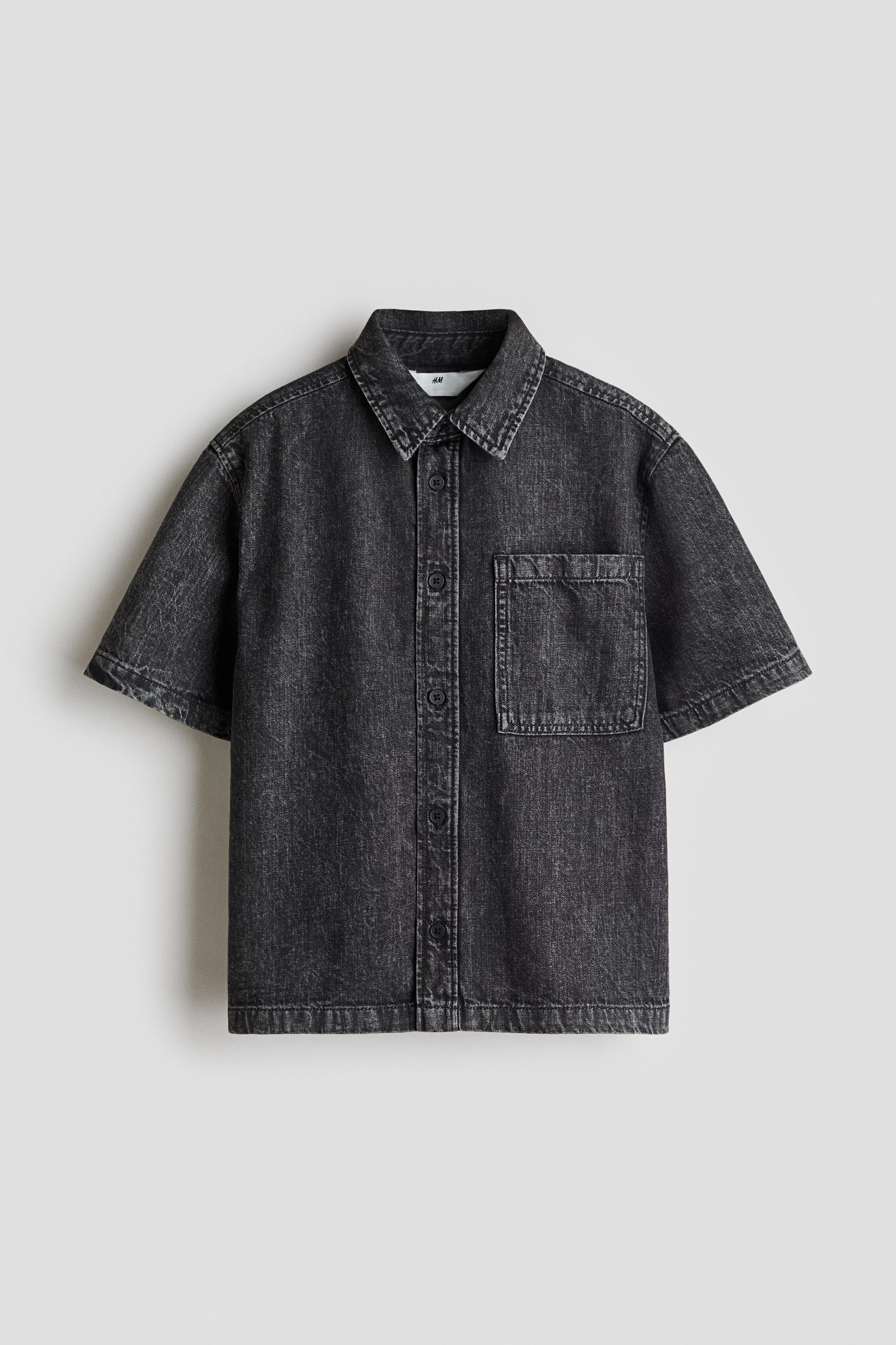 Visualizza immagine più grande: Una camicia a maniche corte e vestibilità ampia in morbido denim di cotone è rivolta frontalmente. Presenta un colletto, bottoni lungo la parte anteriore e una tasca aperta sul petto a sinistra, con l'orlo che presenta un taglio dritto.