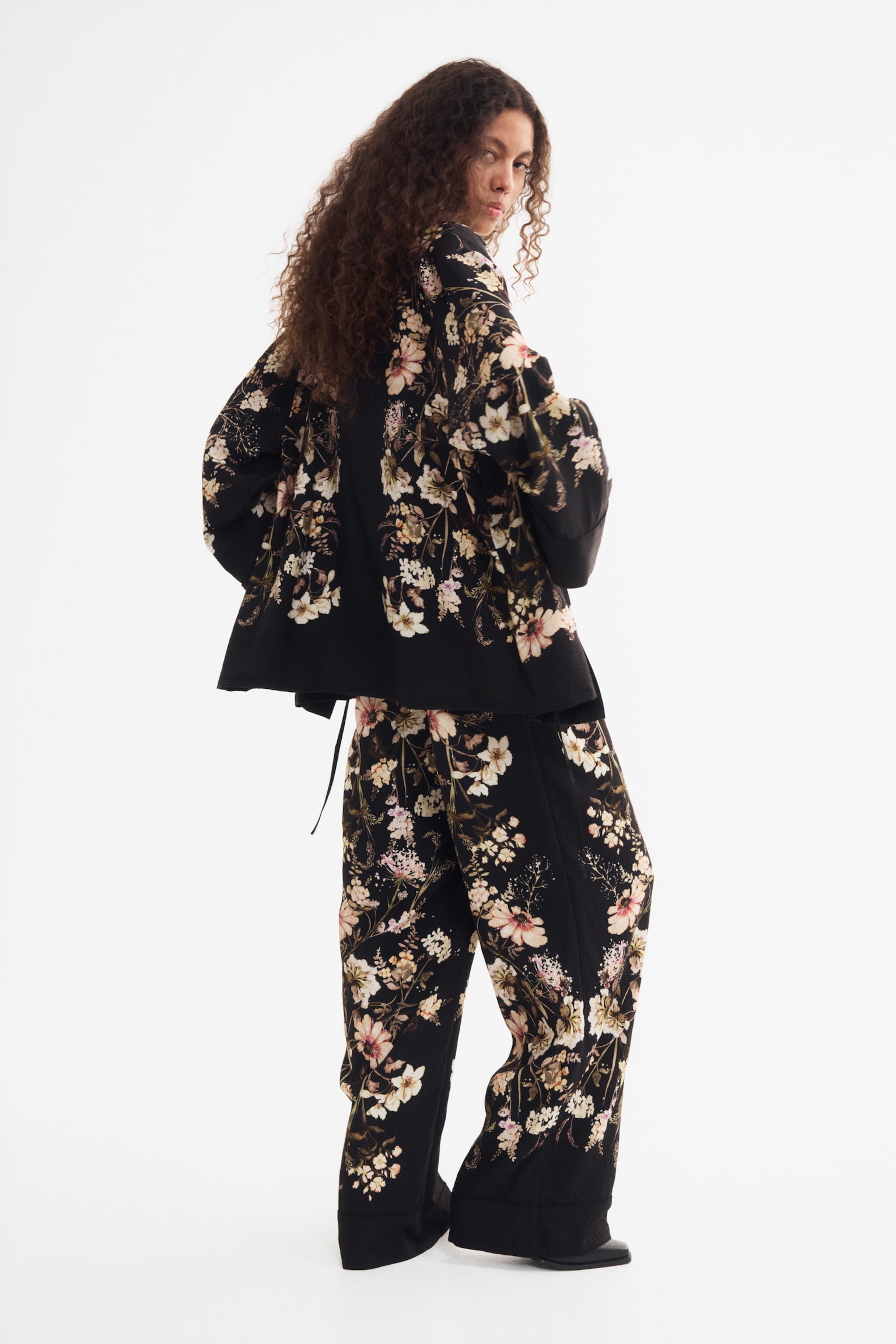 Trousers boast a black base adorned with an intricate, symmetrical floral pattern showcasing light pink, cream, and white blooms, complemented by green and brown foliage, cascading down each wide leg from an elasticated waistband. - 1