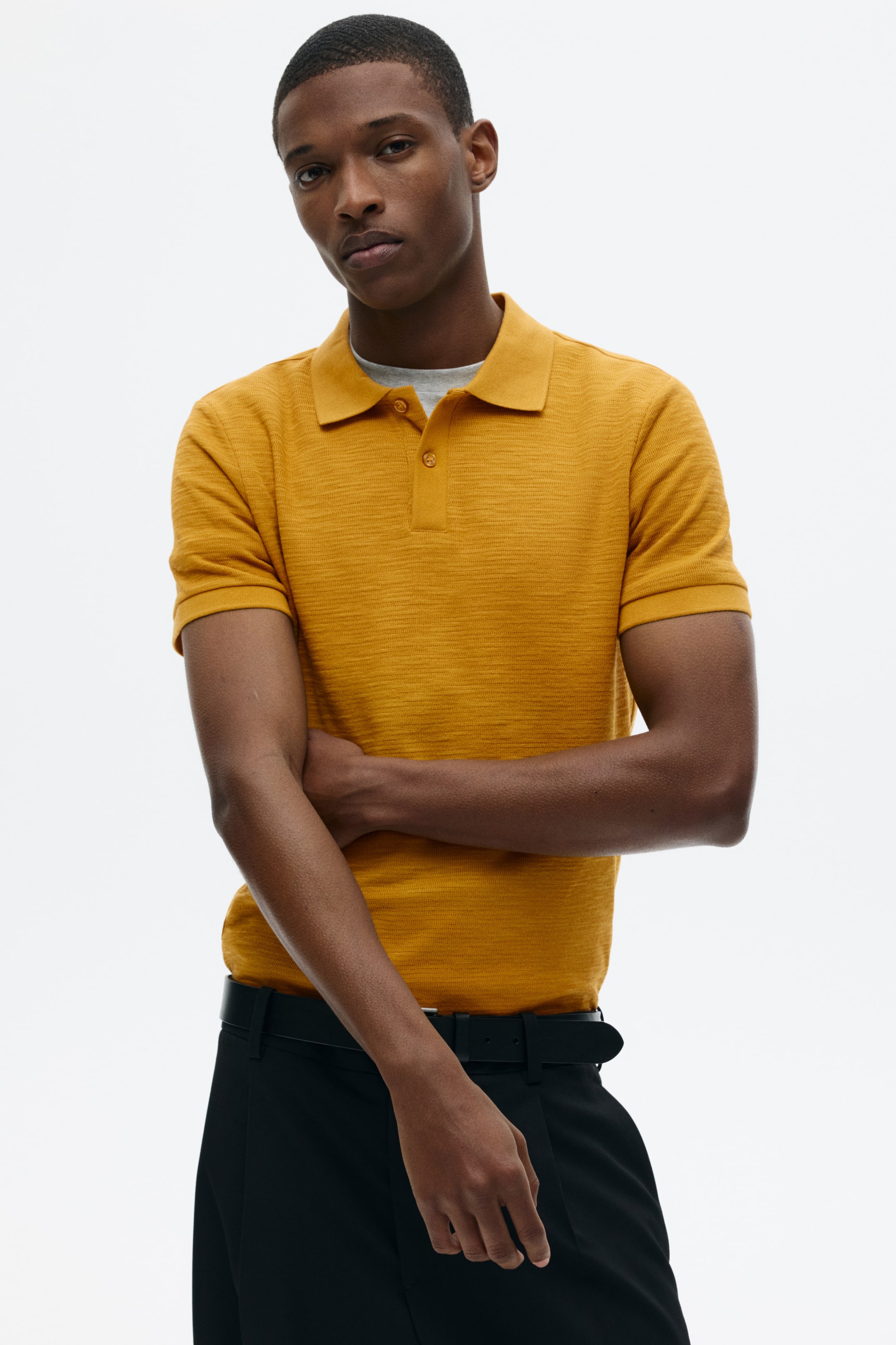 View larger image: A mustard-yellow polo shirt is shown from the front, featuring short sleeves and a subtle horizontal textured knit. It has a two-button placket with matching buttons and a classic spread collar, with a grey crew-neck T-shirt visible beneath the collar.