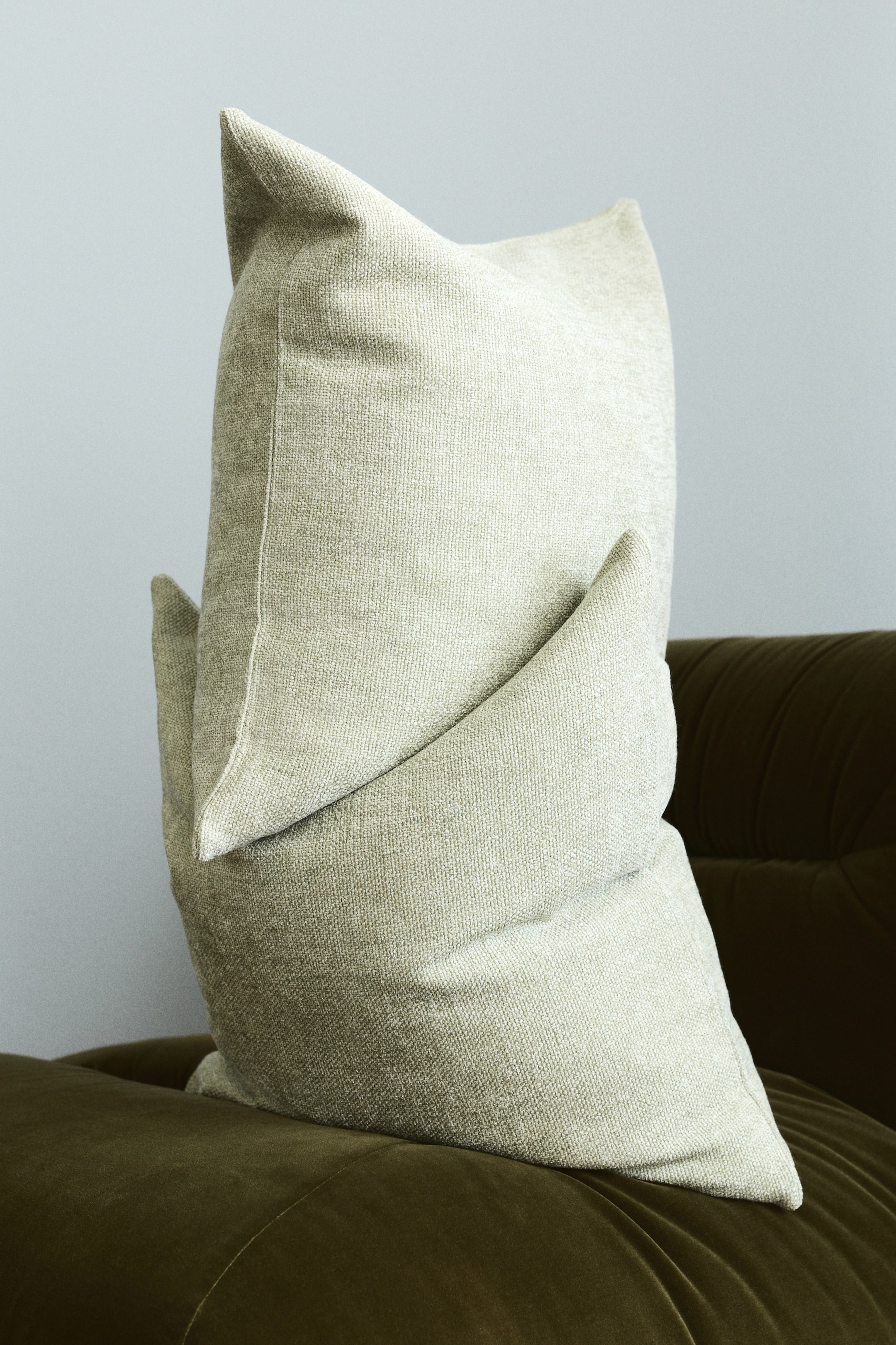 View larger image: A light greige cushion is standing upright, angled slightly to the right, showcasing its front and side. It appears plump with prominent pointed corners, and its subtly textured woven fabric gently creases in the middle.