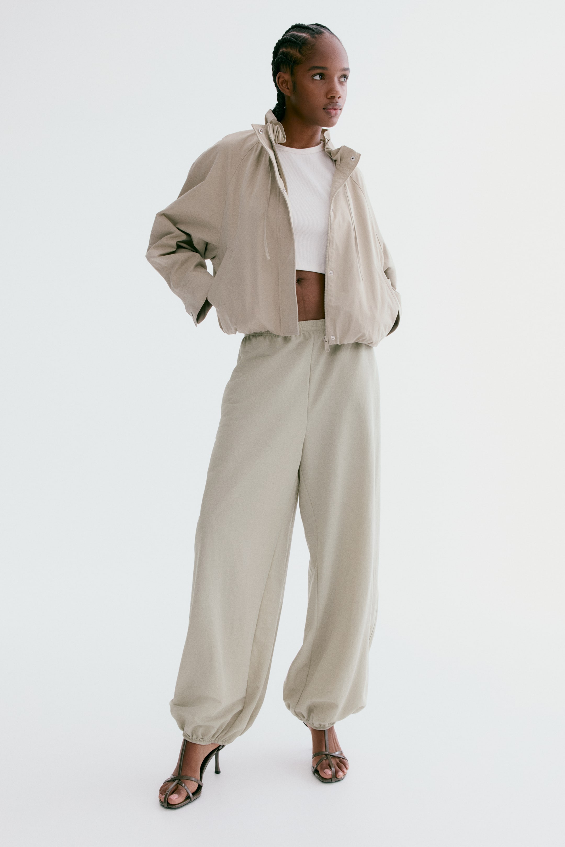Agrandir l'image: Une veste bomber courte beige clair avec un col montant et des manches raglan volumineuses est portée sur un haut court blanc, légèrement orientée vers la gauche. La veste est associée à un pantalon assorti à taille haute et jambes larges, doté d'une taille élastiquée et de chevilles froncées, porté avec des sandales à talons hauts et brides sombres.