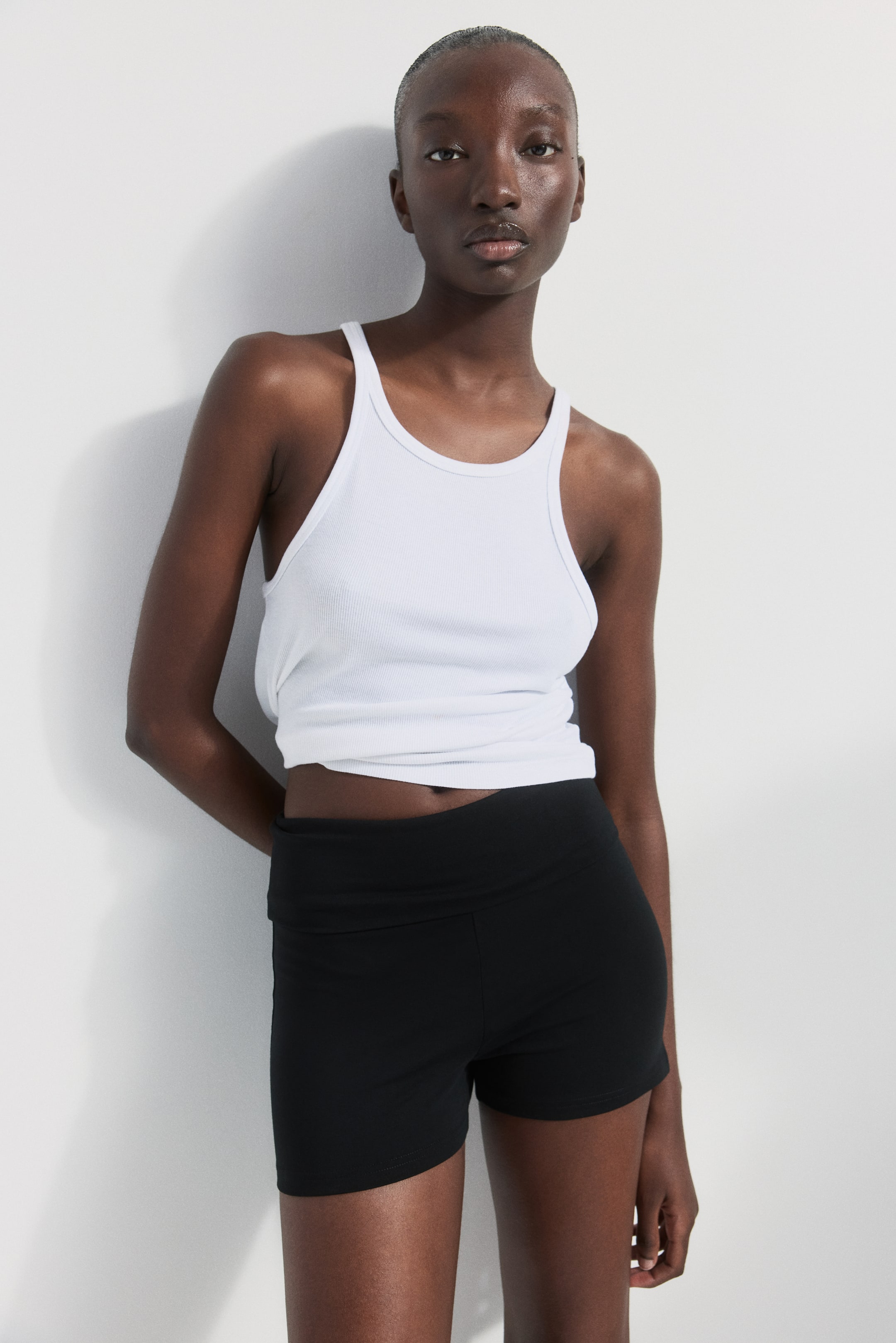 Grösseres Bild ansehen: Die Figur betonenden Hotpants aus schwarzem Stretch-Baumwolljersey mit hoher Taille und breitem Umschlagbund werden vom Model getragen. Sie werden mit einem weißen, gerippten Tanktop kombiniert.