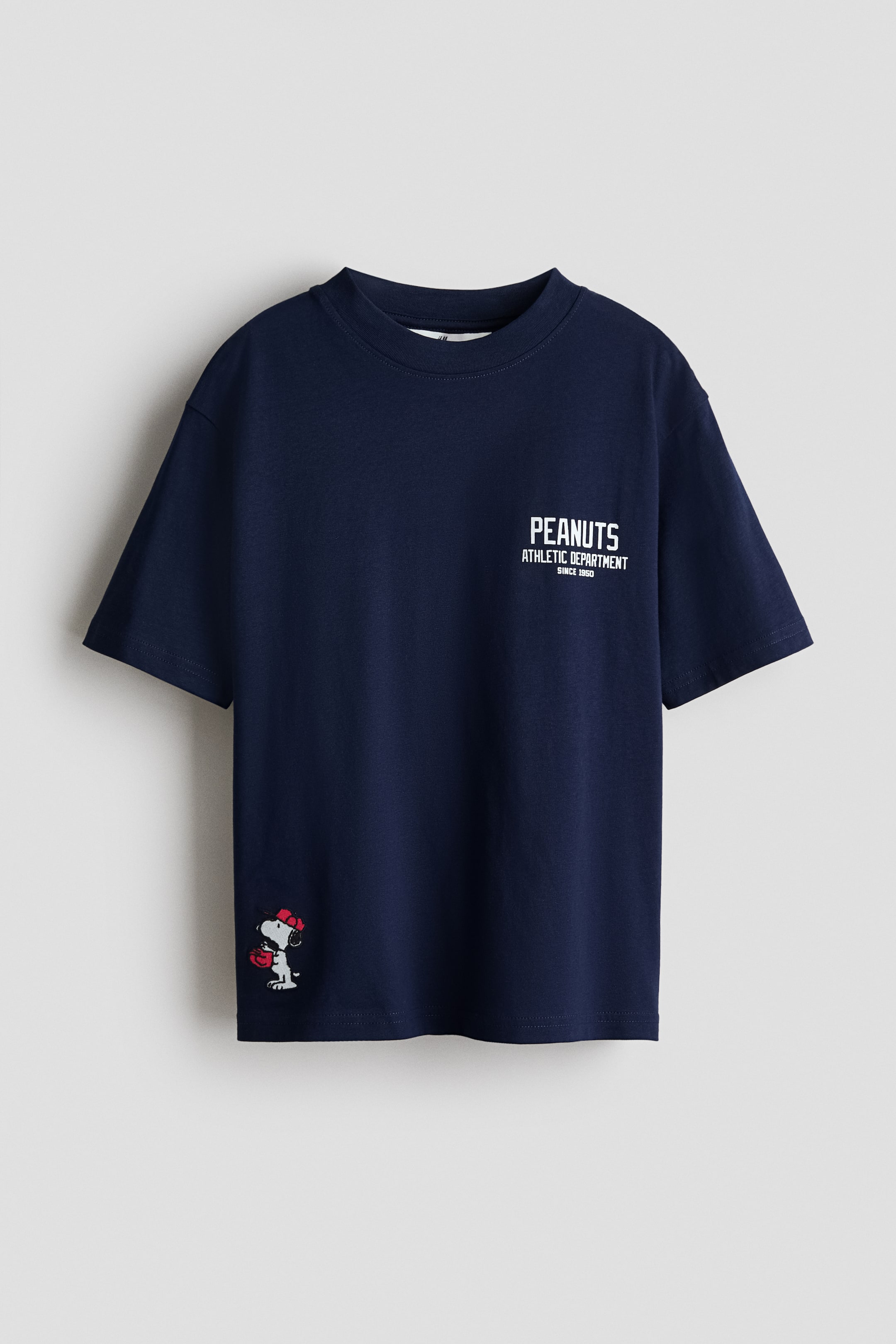 View larger image: A navy blue cotton jersey t-shirt is shown from the front, highlighting its relaxed fit, dropped shoulders, and ribbed neckline trim. The front features white text reading 'PEANUTS ATHLETIC DEPARTMENT SINCE 1950' on the left chest, along with a small Snoopy graphic wearing a red cap and red gloves on the lower left.