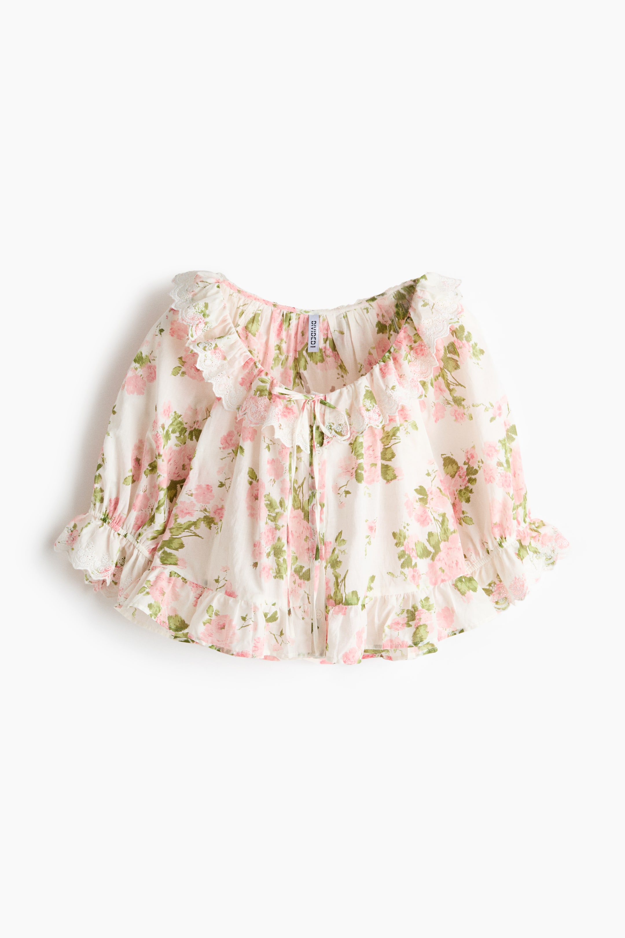 View larger image: The blouse is displayed from the front, showcasing a floral design with pink roses and green leaves set against a light backdrop. The deep, round neckline is embellished with scalloped flounce and broderie anglaise, complemented by narrow ties. The elbow-length sleeves come with elasticated cuffs. Designed with a gathered waist and a flounced hem, this blouse offers a perfect blend of style and comfort.