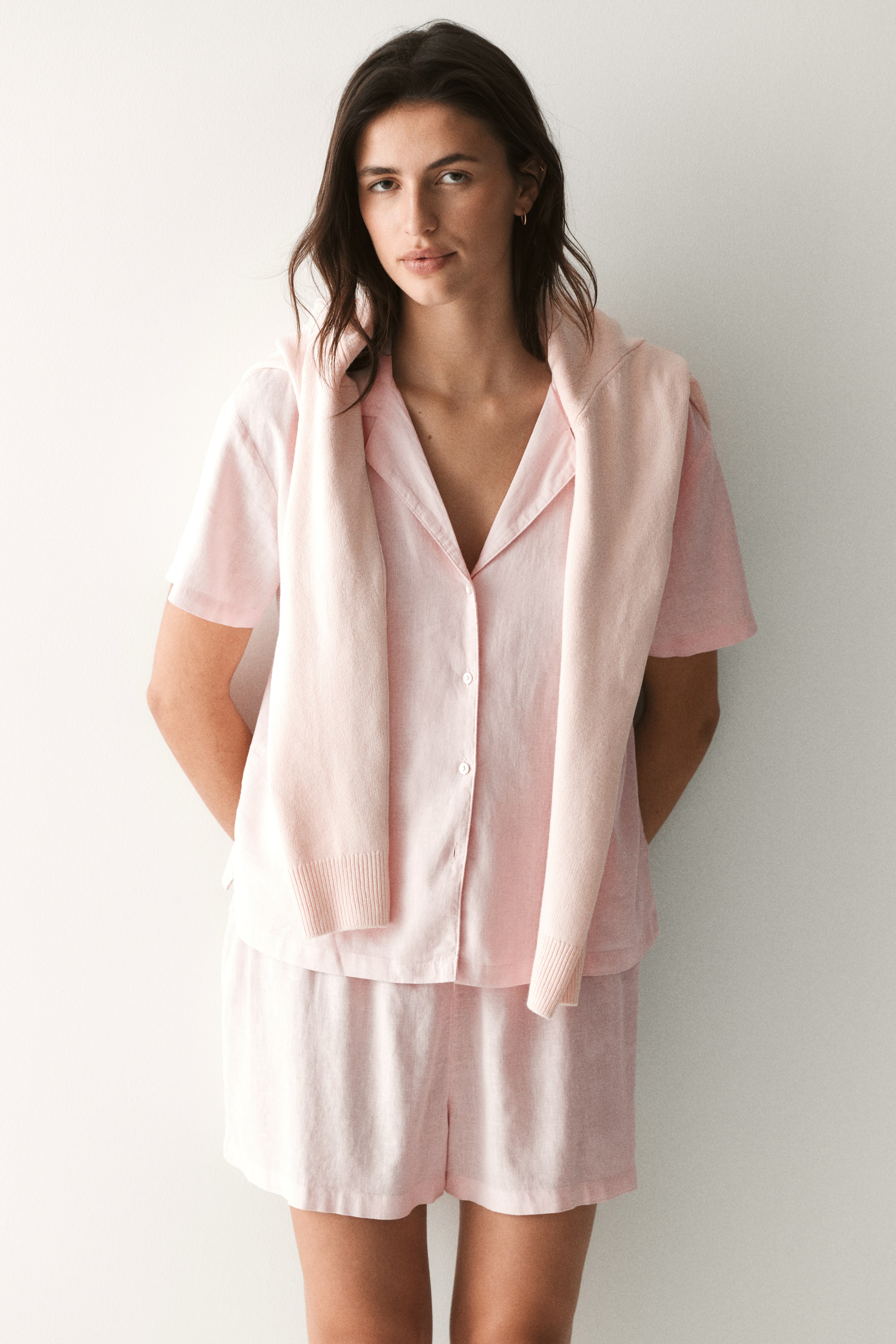 View larger image: The model is showcasing a breezy pink pyjama set, featuring a short-sleeved shirt with a resort collar and button-down front, paired with matching shorts that have an elasticated waist. Draped over the model's shoulders is a pink knitted sweater.