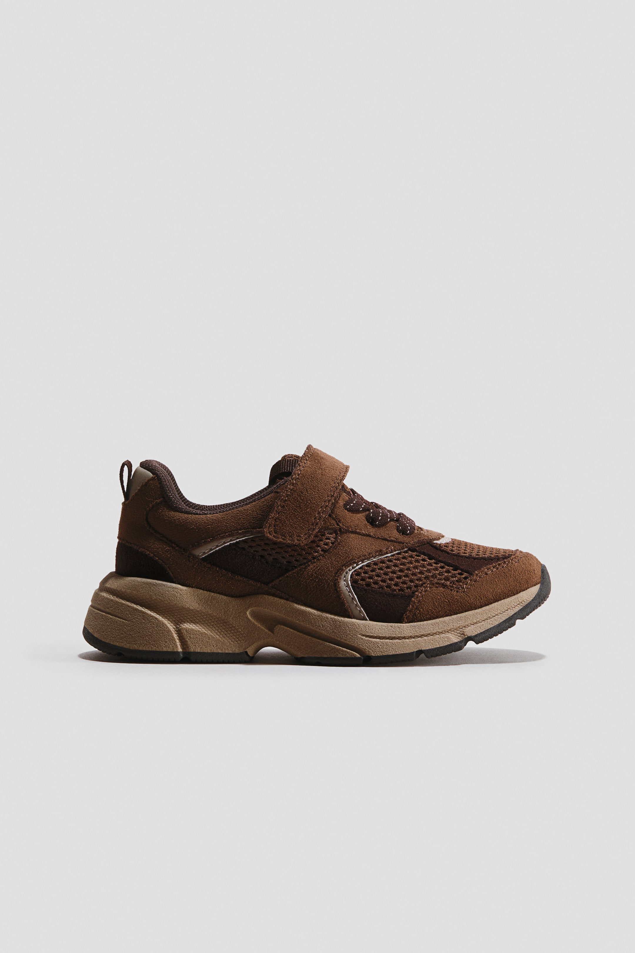 View larger image: A brown sneaker is showcasing its left side profile, adorned with dark brown suede-like overlays and lighter brown mesh panels. It features a single velcro strap over elastic laces and a chunky, multi-layered sole in shades of brown and tan.