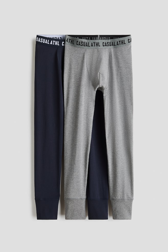 Two pairs of men's thermal leggings, one in navy blue and one in heather grey, are hanging flat in a front-facing view. Both pairs feature a wide elastic waistband printed with the repeated text "CASUAL ATHL" and finish with ribbed ankle cuffs. - 1