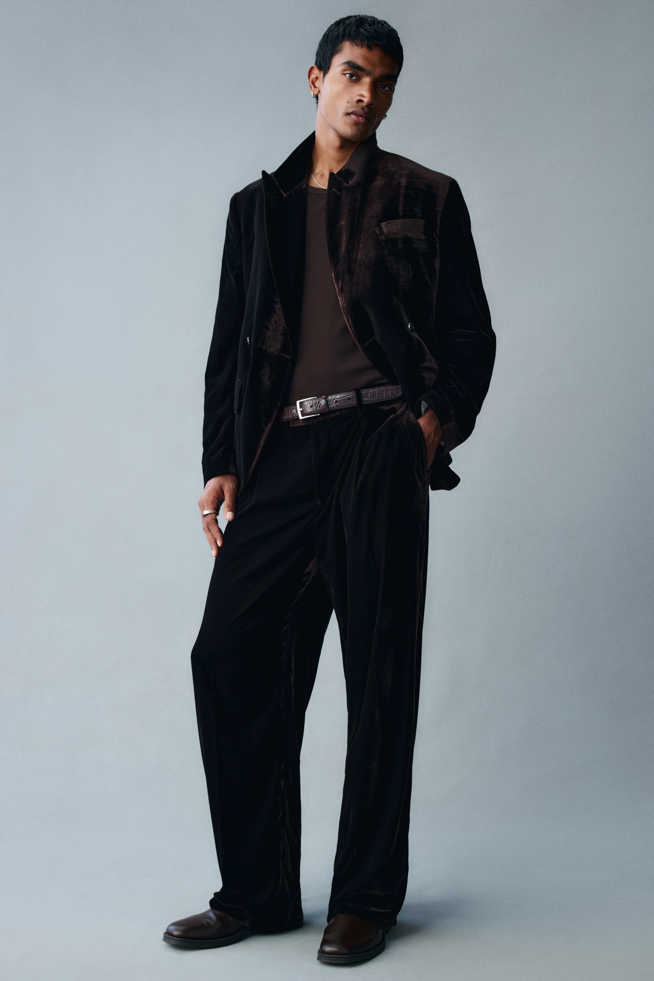 View larger image: Relaxed-fit velvet trousers in a dark brown shade are being worn by the model, who is standing with one hand resting in the side pocket. The trousers feature a straight-leg silhouette with front creases and are styled with a matching velvet blazer, a textured brown belt, and dark leather shoes.