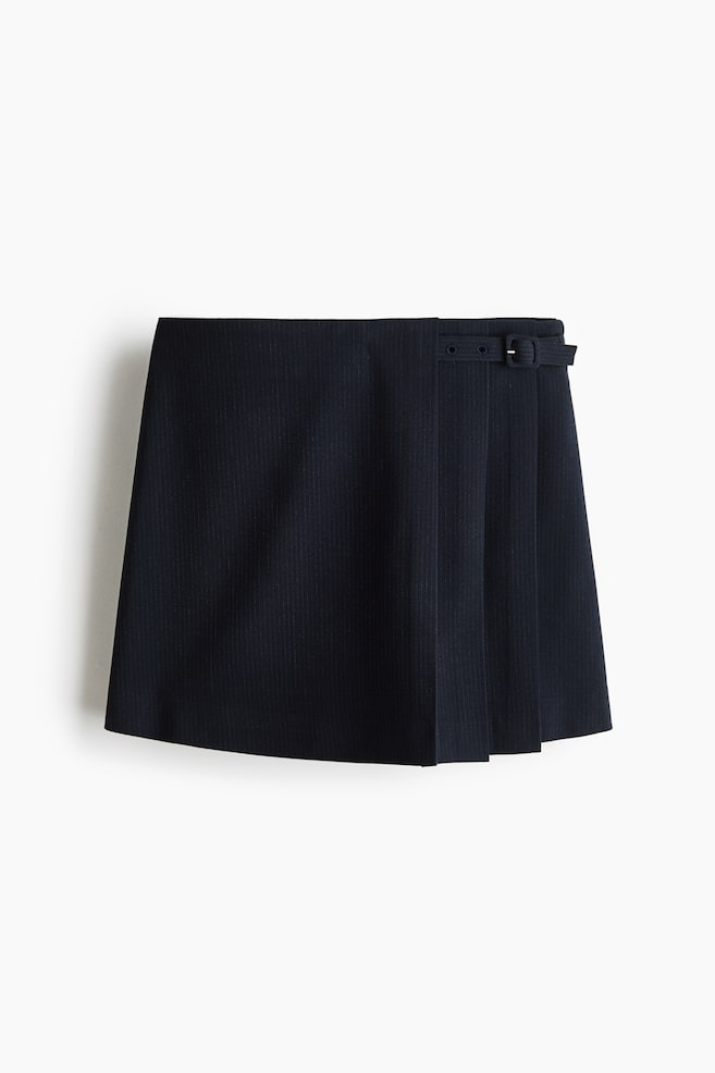 Se muestra una minifalda pantalón de rayas diplomáticas en azul marino oscuro desde el frente, con diseño de talle alto cruzado y pliegues verticales en un lado. Un cinturón decorativo a juego con hebilla forrada detalla la cintura en el lado plisado. - 1