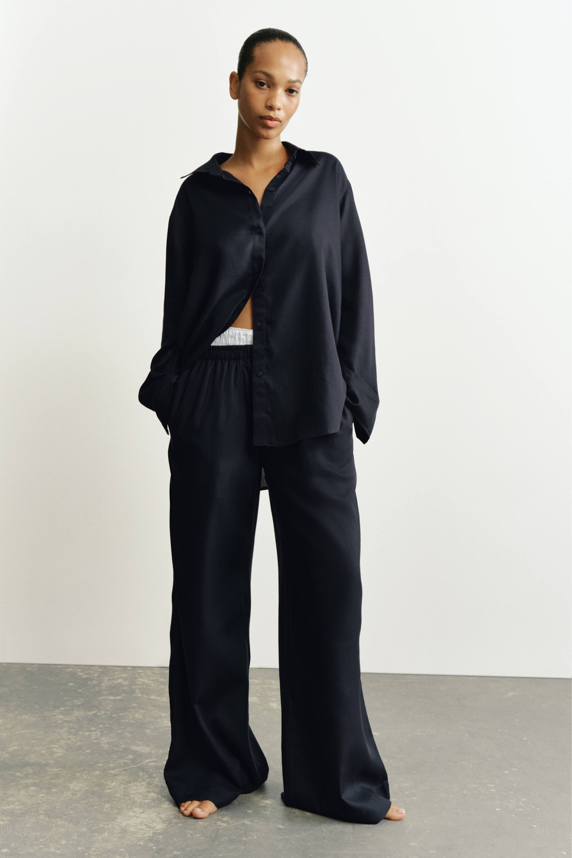 Größeres Bild anzeigen: Die Hose aus dunklem Lyocell-Gewebe mit entspannter, weit geschnittener Silhouette und elastischem Bund wird vom Model getragen. Das Model steht mit den Händen in den Seitentaschen und kombiniert die Hose mit einem passenden, aufgeknöpften Langarmhemd, das einen weißen Bund freilegt.
