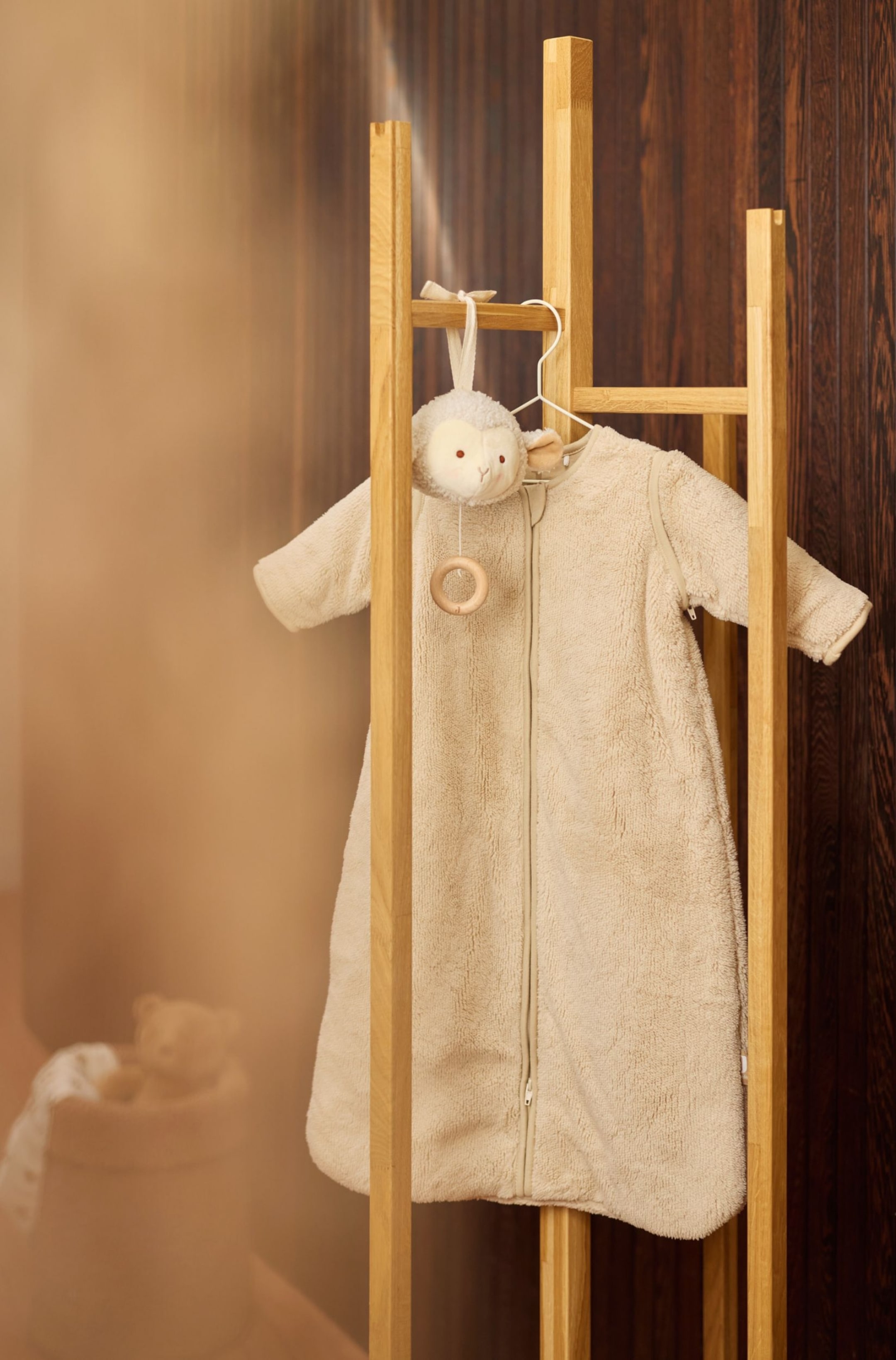 Agrandir l'image: Une gigoteuse bébé en polaire pelucheuse beige est suspendue de face sur un support en bois, avec une fermeture éclair frontale sur toute la longueur et de longues manches amovibles zippées aux coutures d’épaules. Un petit jouet mouton en peluche avec un anneau en bois est suspendu au crochet du cintre juste au-dessus du col.