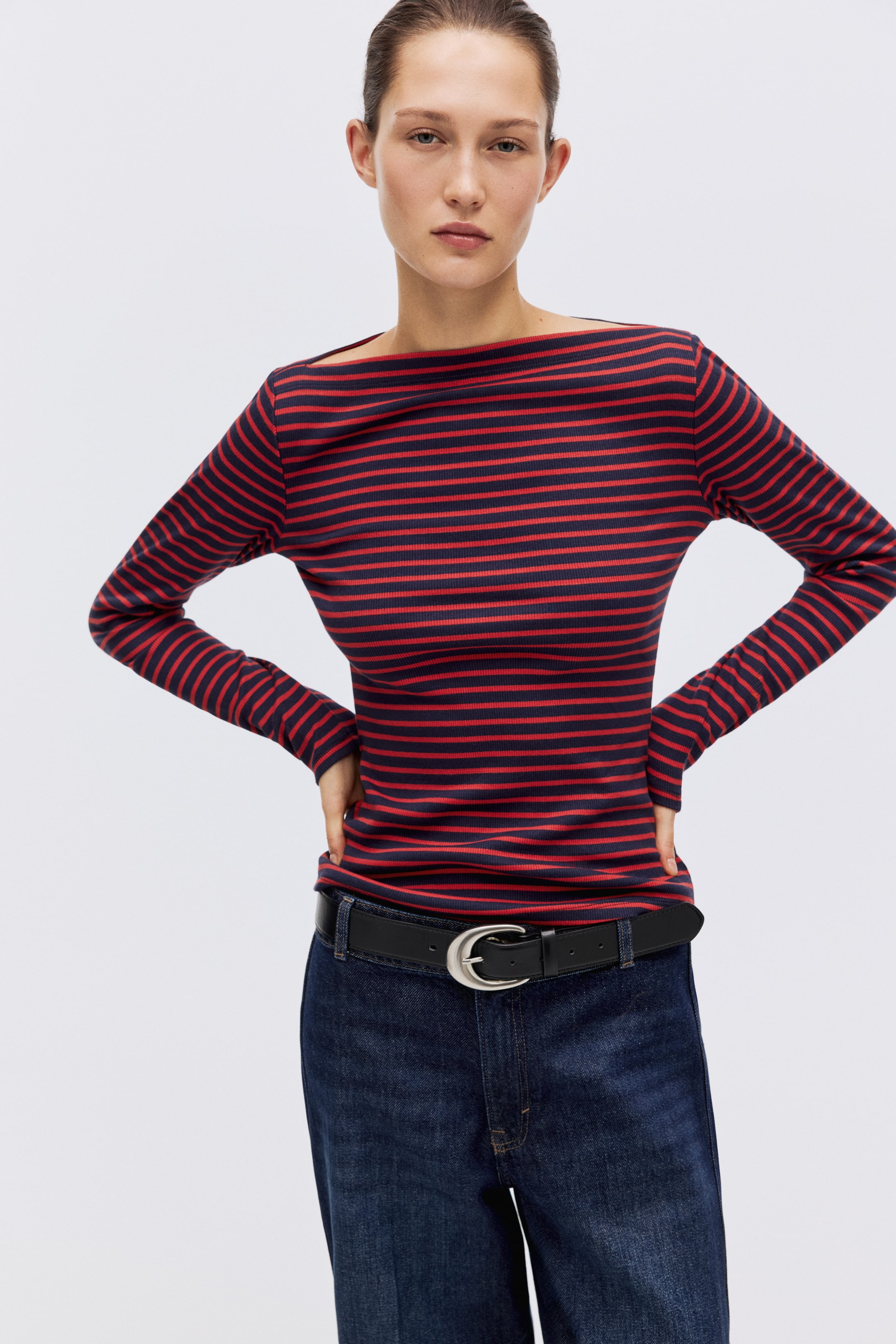 View larger image: A striped long-sleeve top featuring close-set red and navy horizontal stripes on ribbed fabric. It presents a boat neckline and a slim fit, and is tucked into dark-wash jeans.