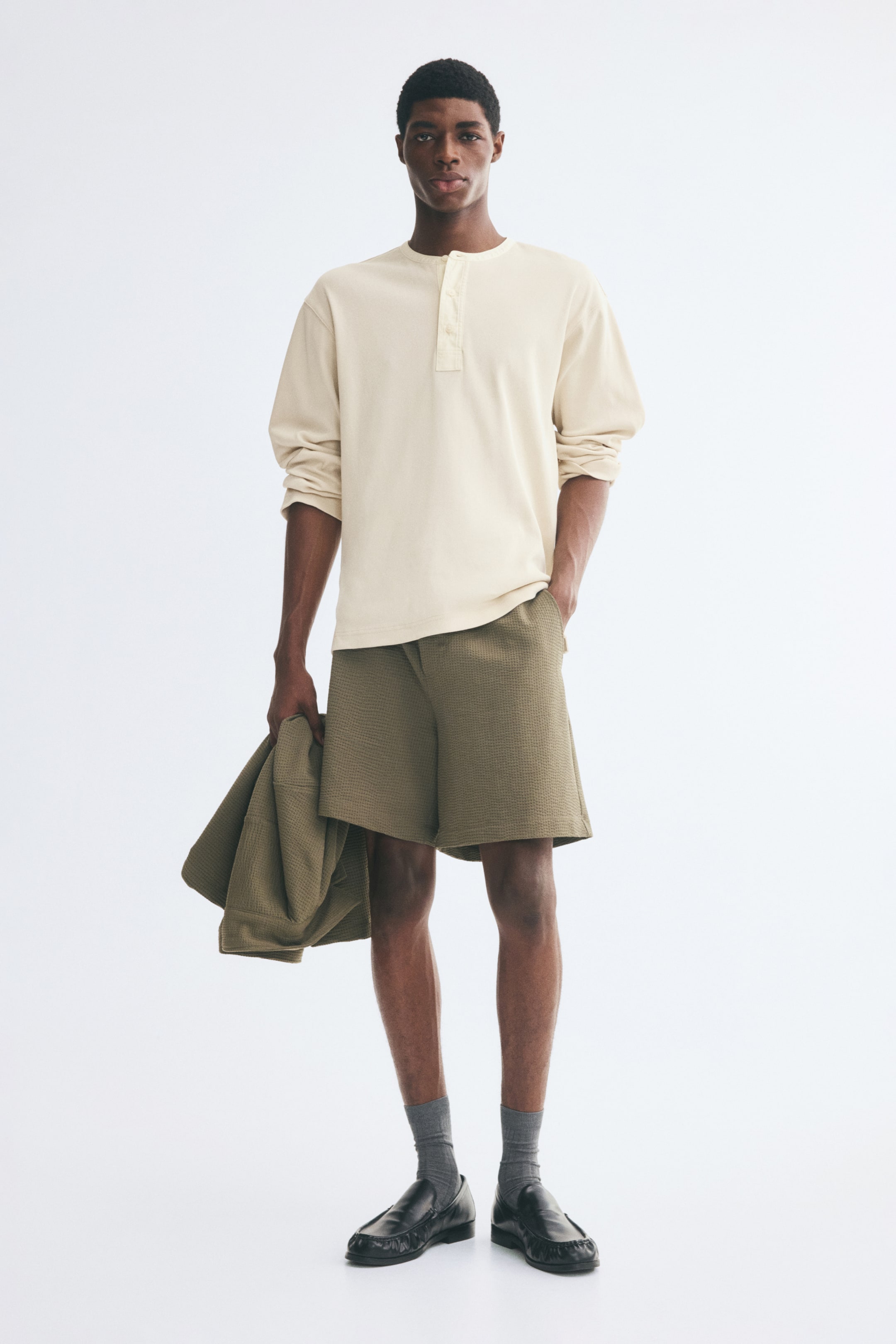 View larger image: The model is wearing mid-length shorts in an olive green waffled jersey fabric, standing with one hand placed in a diagonal side pocket. The relaxed-fit shorts are paired with a cream-coloured long-sleeve henley shirt, grey socks, black loafers, and a matching waffled jacket held in the model’s other hand.