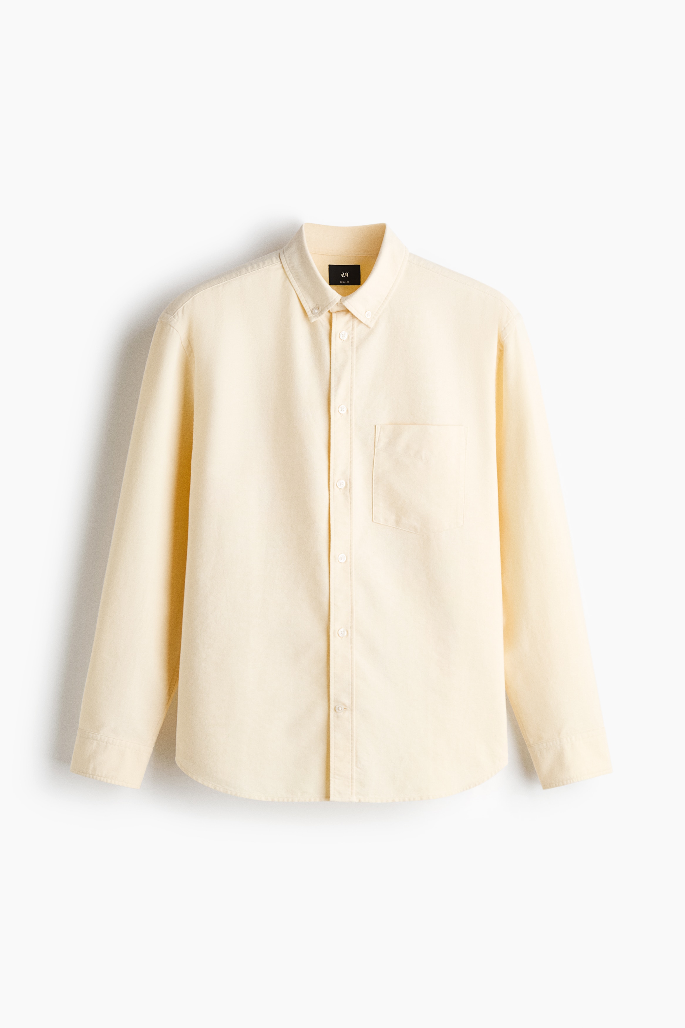 The model is wearing a pale yellow Oxford cotton shirt layered over a white T-shirt and tucked into dark trousers. The shirt features a button-down collar, a classic button-front closure, a single chest pocket, and long sleeves rolled up at the cuffs.