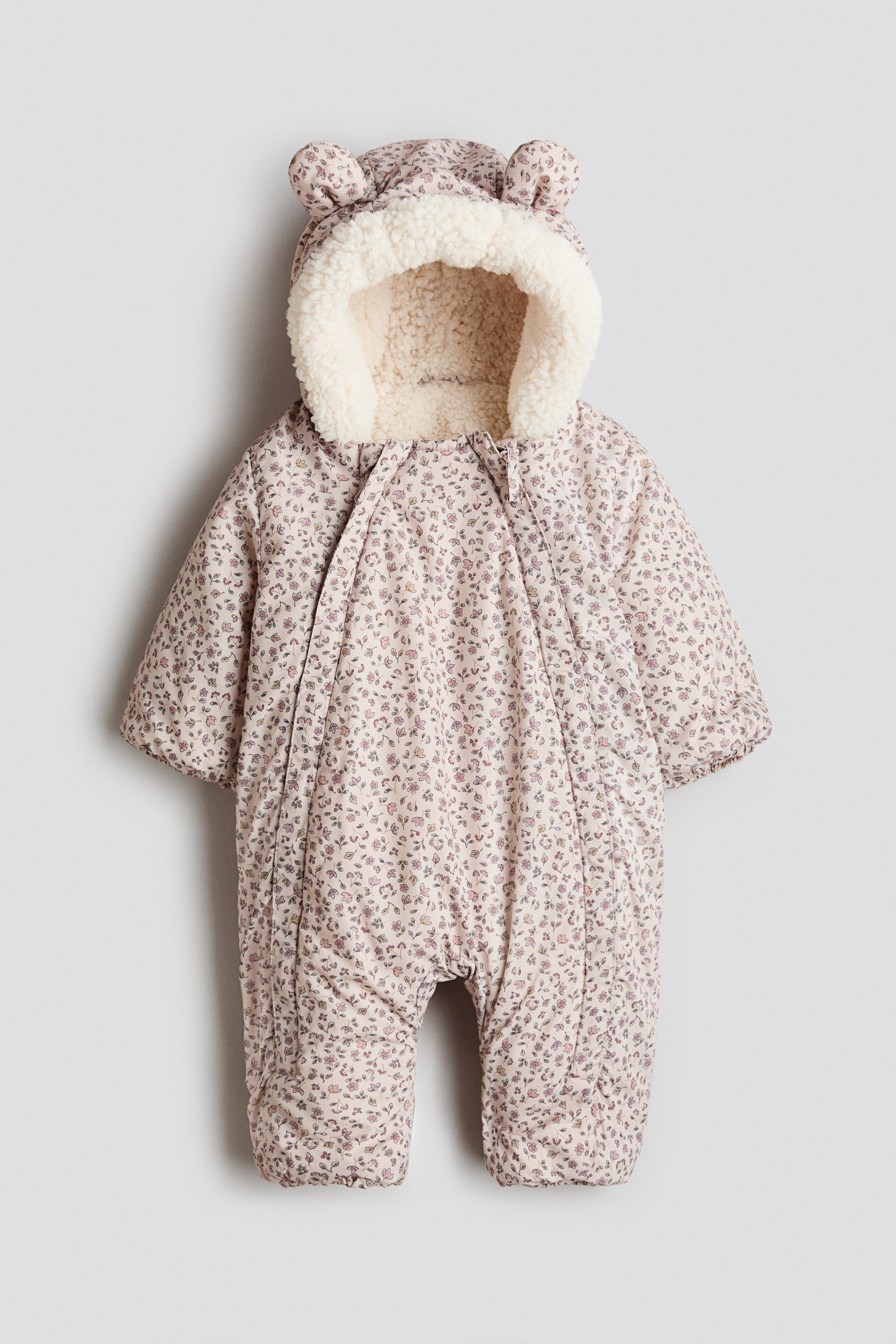 Visa större bild: En vadderad snöoverall för baby sedd framifrån visar en ljusrosa bas med ett litet blommönster. Den har en integrerad huva med björnöron och ett sherpaliknande foder i krämfärg, samt elastiska muddkanter och öppningar vid anklarna.