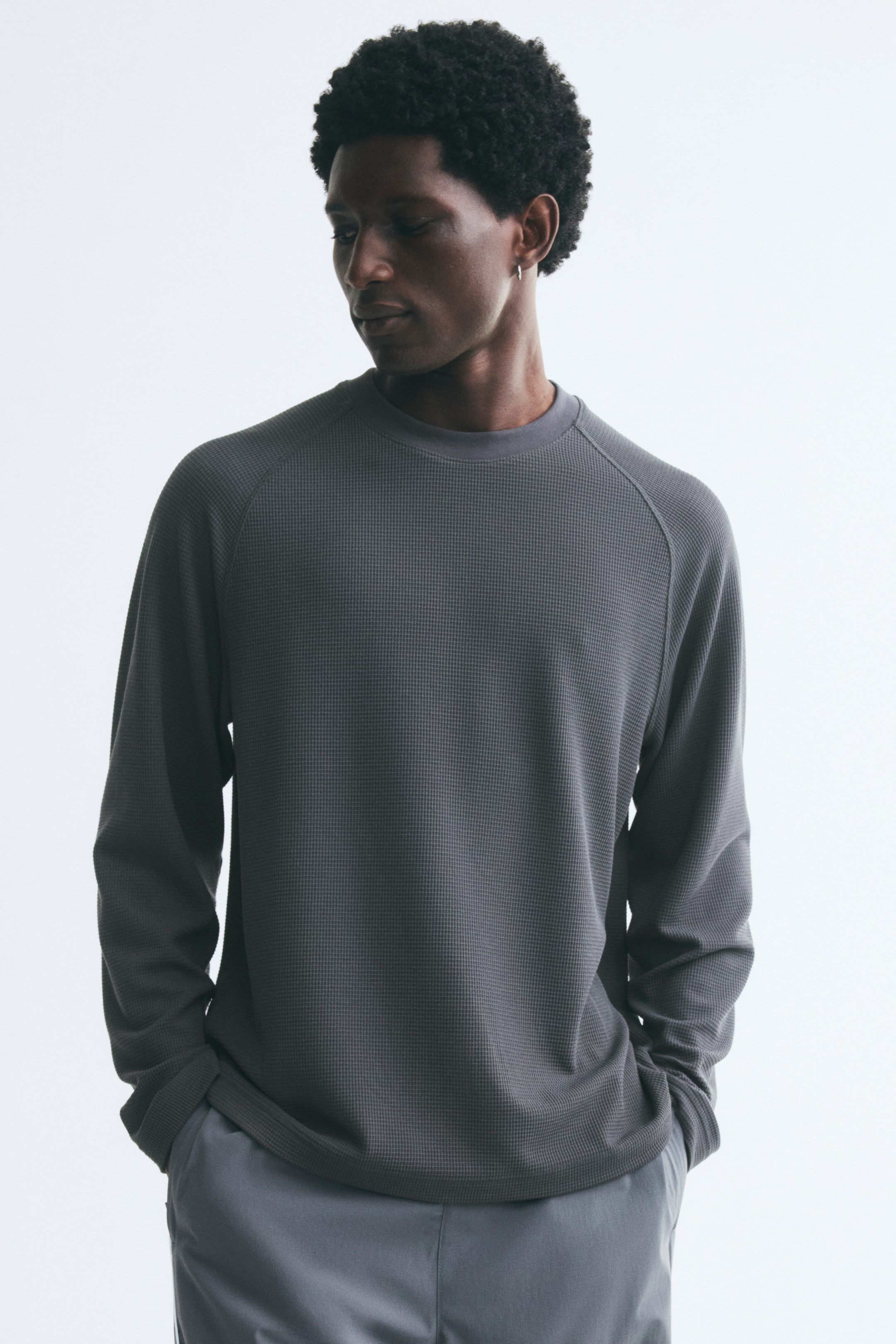Voir une image plus grande: Le mannequin porte un tee à manches longues gris foncé en jersey gaufré, à coupe décontractée, avec un col rond et des manches raglan. Le mannequin est debout, les mains dans les poches d’un pantalon gris pâle, mettant en valeur l’ourlet droit du haut.