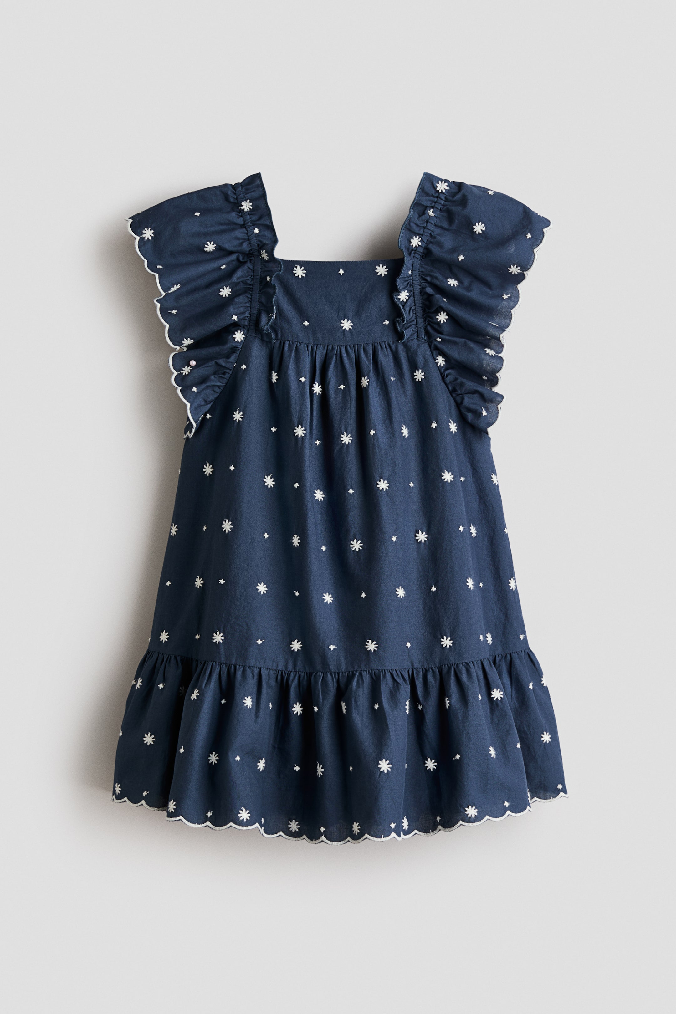 Visa större bild: En mörkblå klänning med fyrkantig urringning, volangprydda ärmar med vit kant och vita broderade stjärnliknande motiv utspridda över framsidan. Klänningen har ett rynkat lager vid midjan och en volangkant, även avslutad med en vit kant.