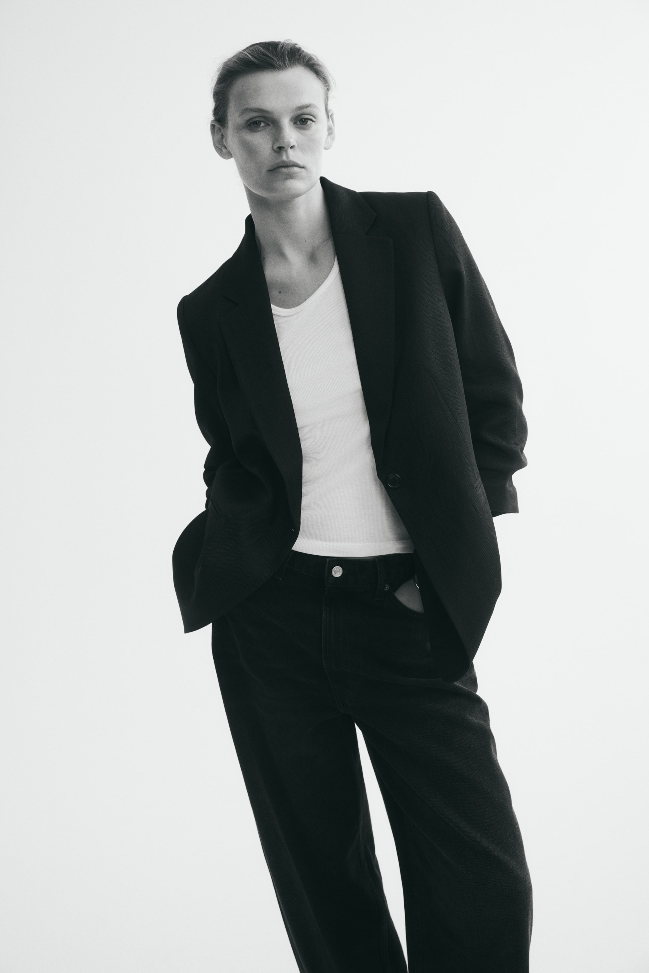 Voir une image plus grande: Un long blazer noir à simple boutonnage, avec revers crantés, épaules structurées et manches trois-quarts froncées, est porté par le modèle. Le blazer non boutonné est superposé à un débardeur blanc et agencé avec un pantalon foncé.