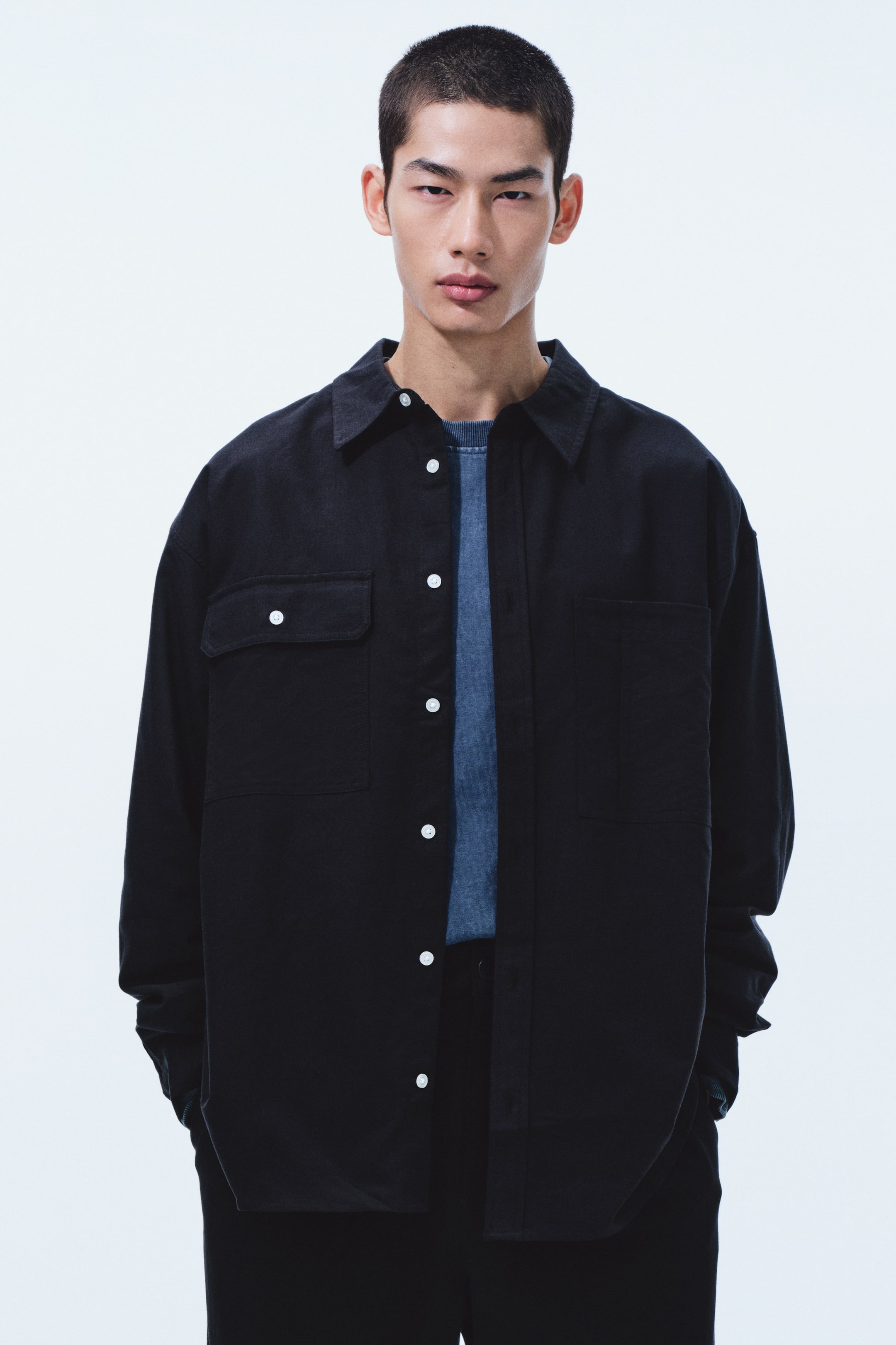 拡大画像を表示: 黒のオーバーサイズのオーバーシャツを、青のテクスチャーのあるクルーネックトップの上に開けて羽織っており、先の尖った襟、白いボタンの前開き、胸にフラップ付きポケットが2つ付いています。