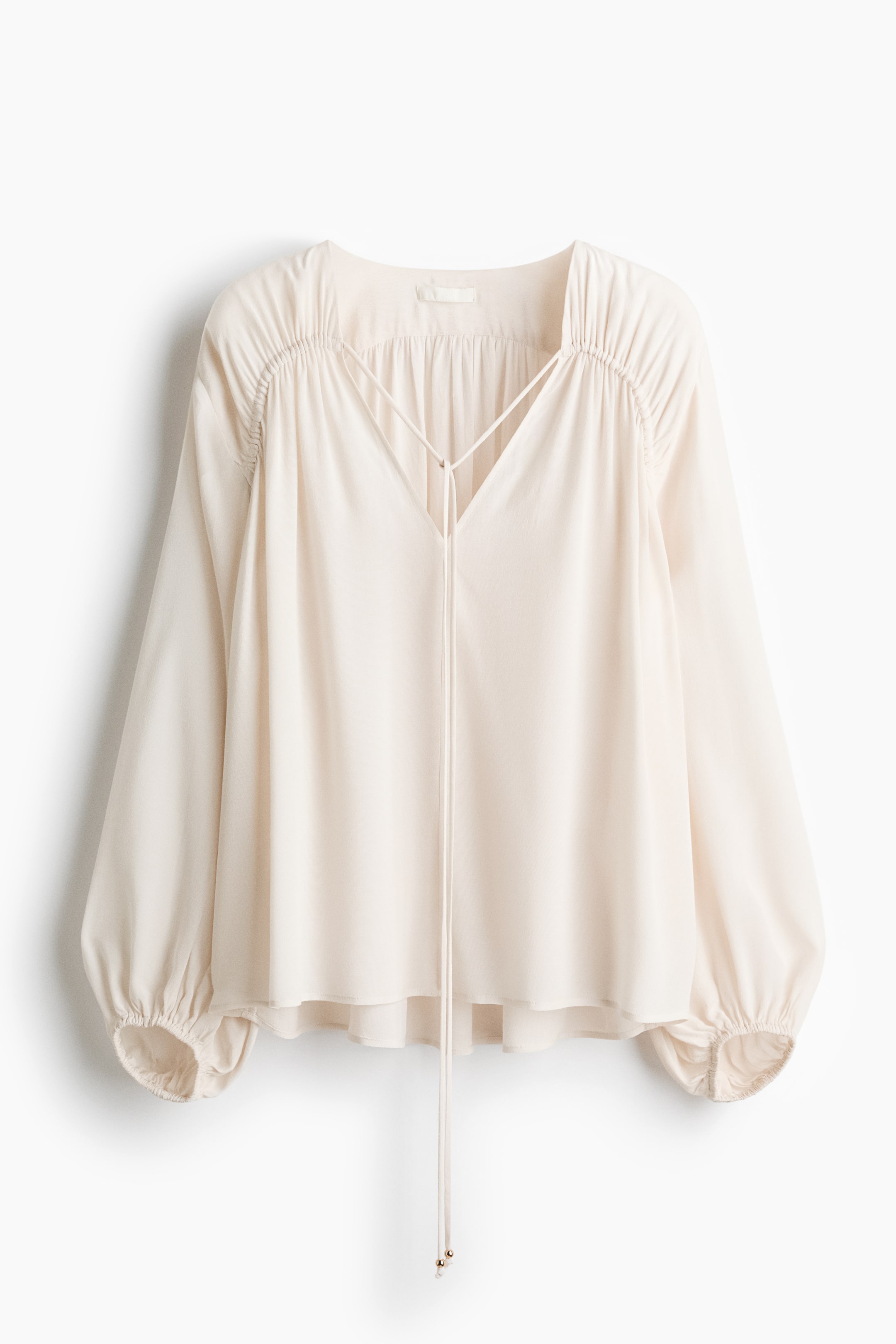 View larger image: FRONT view of a cream-coloured blouse, featuring a V-neckline with a tie closure. It is displaying long, voluminous sleeves that are gathered at the shoulders and cinched at the cuffs, and is presenting a loose-fitting silhouette.