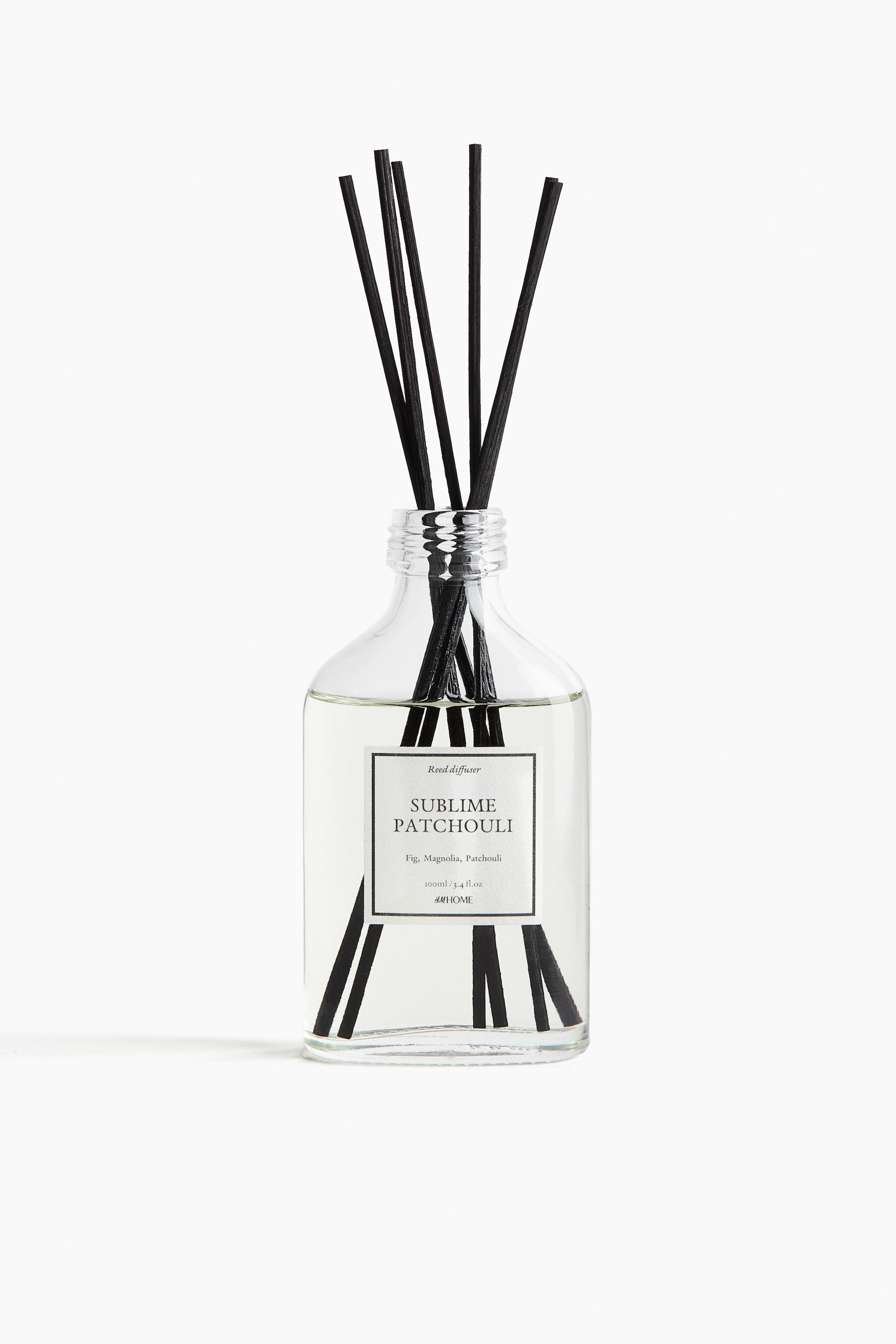 View larger image: A reed diffuser is presented from the front, featuring a clear glass bottle with a threaded neck holding clear liquid. Several black reeds are standing upright, partially submerged in the liquid and extending from the bottle's neck, while a white rectangular label with "Sublime Patchouli" is affixed to the front.
