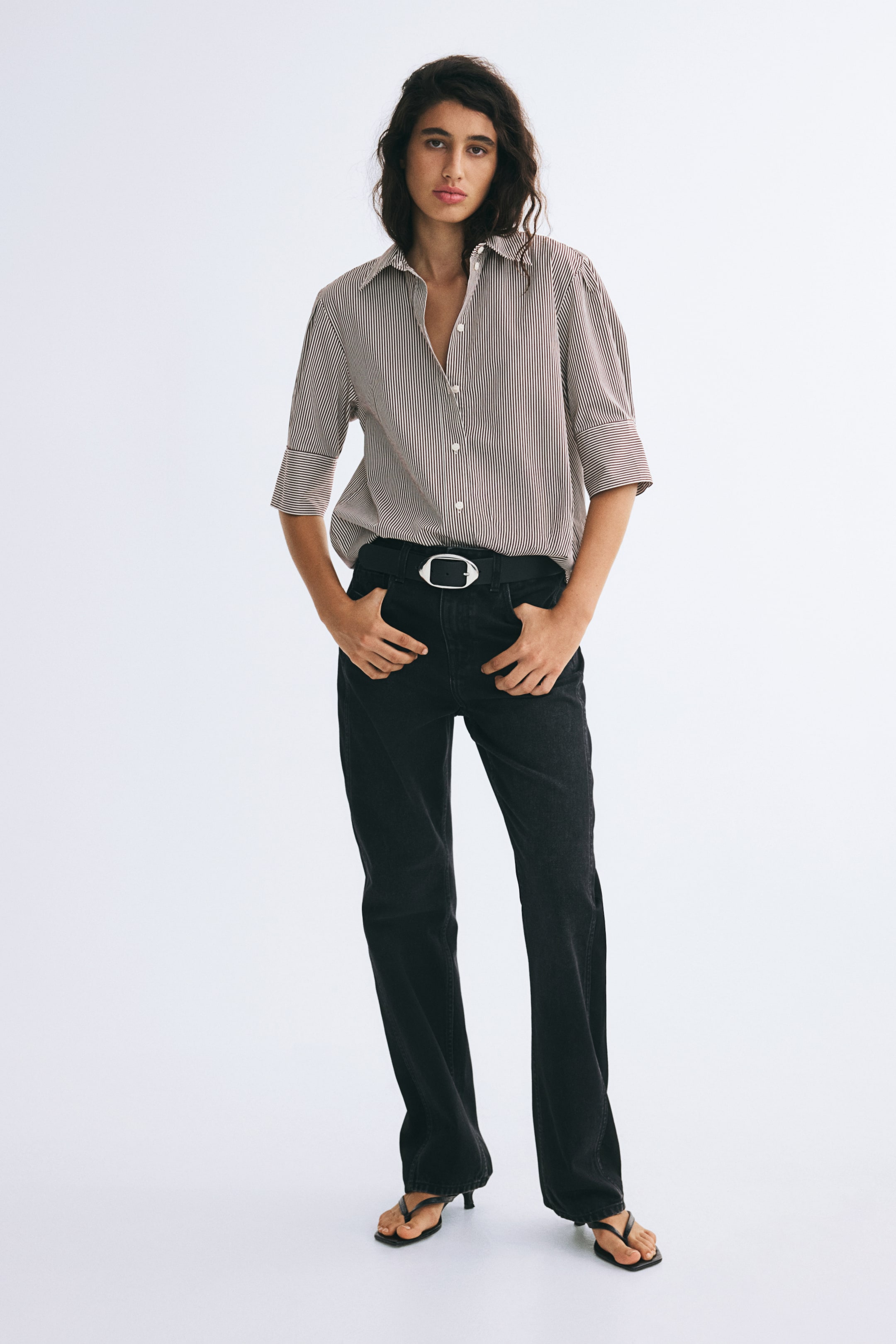 View larger image: A striped cotton poplin shirt featuring a collar, a button-down front, and pleated, elbow-length sleeves with wide cuffs is being worn by the model. The shirt is tucked into black jeans styled with a black belt.