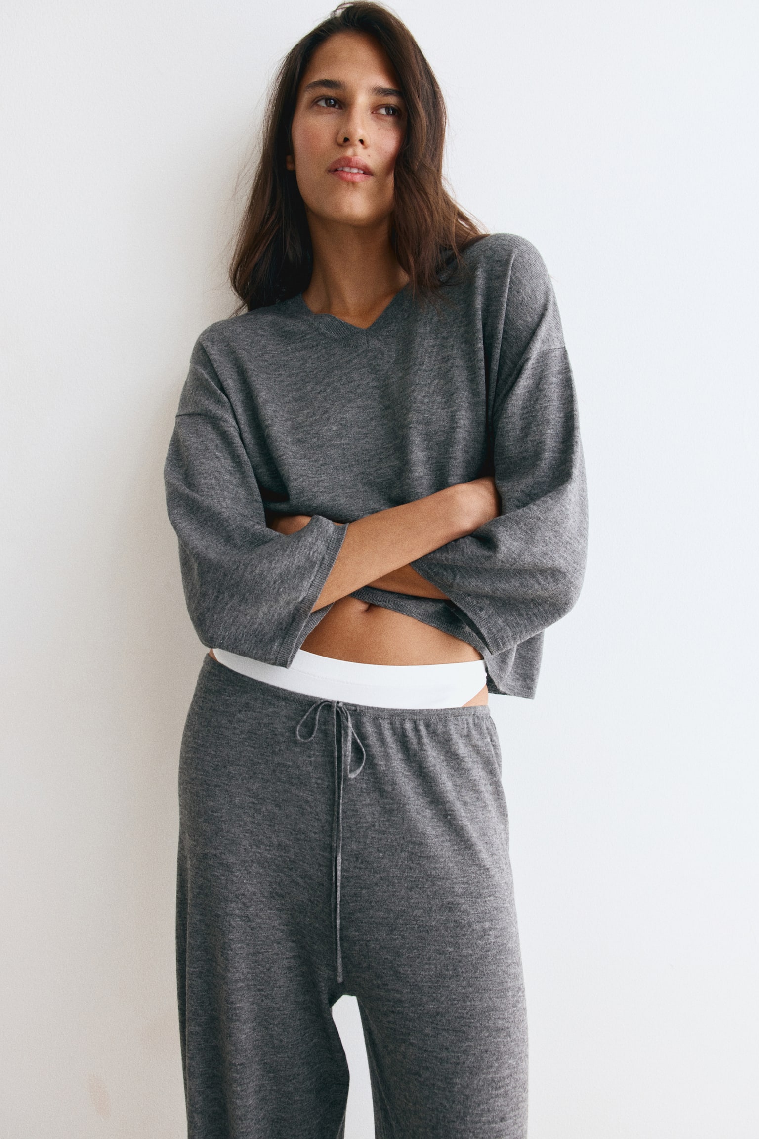 A grey wool-blend knit sweater featuring a V-neck, dropped shoulders, and wide 3/4-length sleeves is being worn by the model with crossed arms. The sweater is styled with matching grey knit drawstring trousers. - 1