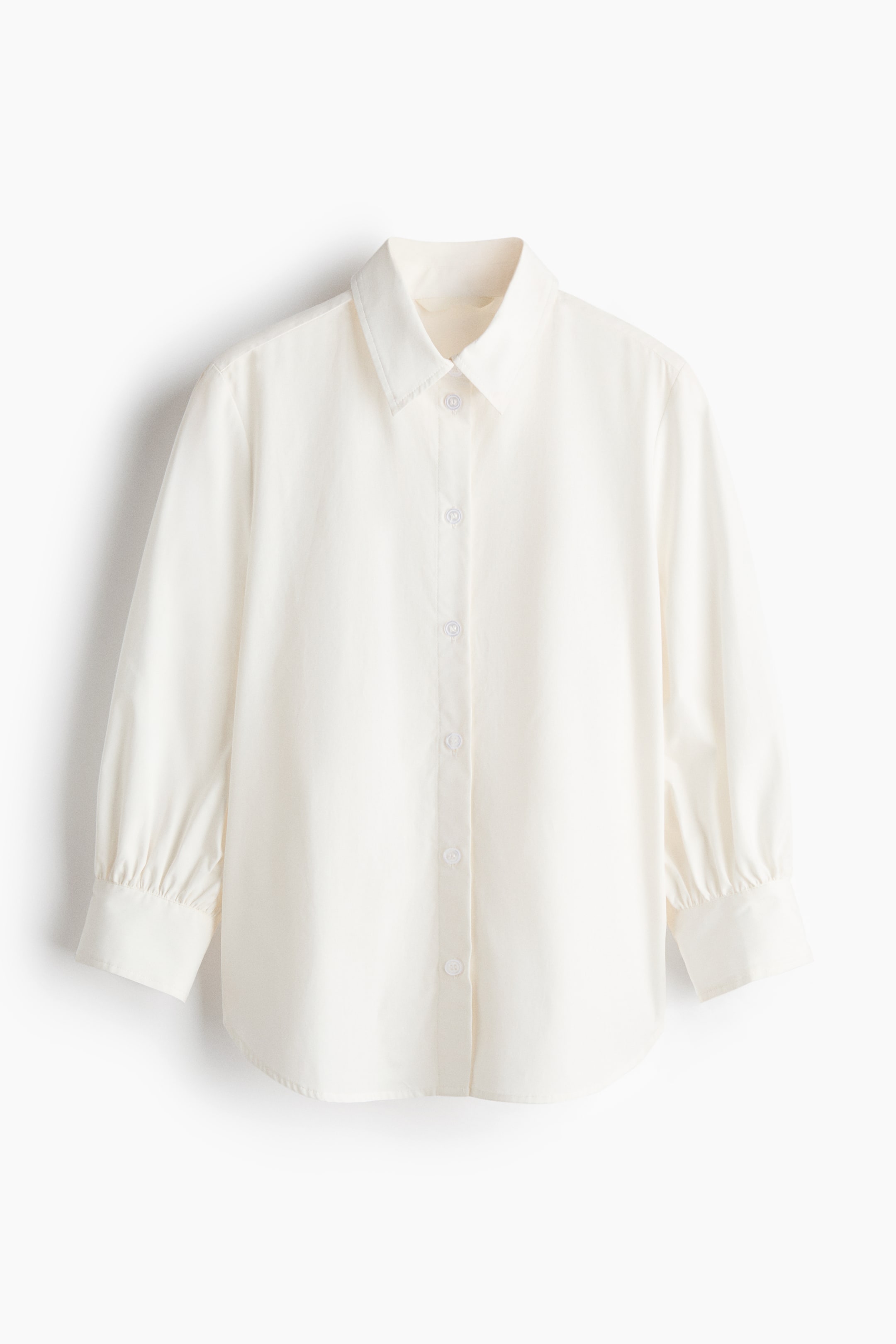 View larger image: A cream shirt is showcasing its front, highlighting a pointed collar, a full-button placket with white buttons, and long sleeves that gather into single-button cuffs, with its hemline curving gently.