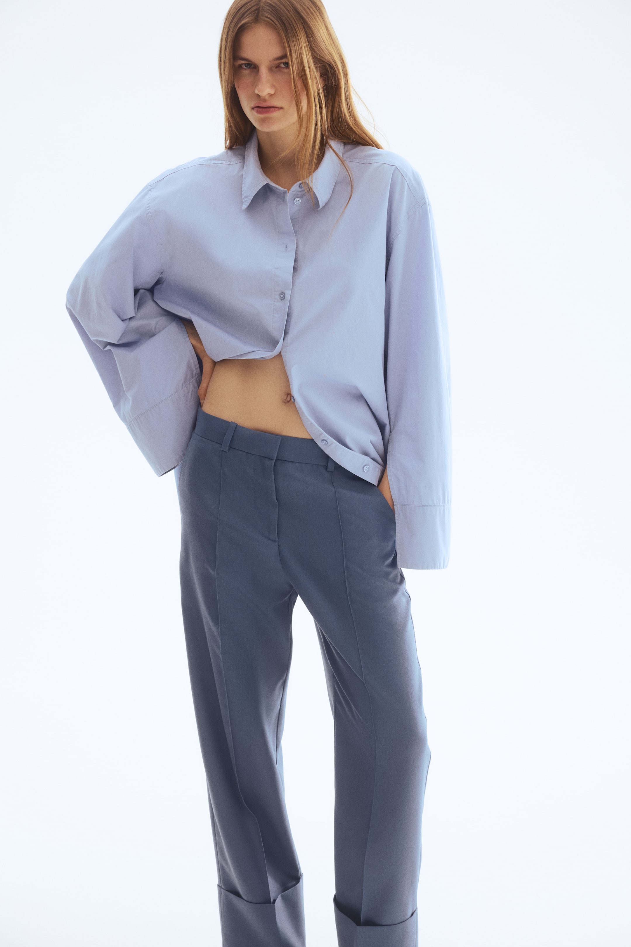 查看大圖: 模特穿著剪裁合身的藍灰色直筒長褲，前面有縫製的折痕，斜插口袋，褲腳有寬闊的翻邊。模特將一隻手放在斜插口袋中。