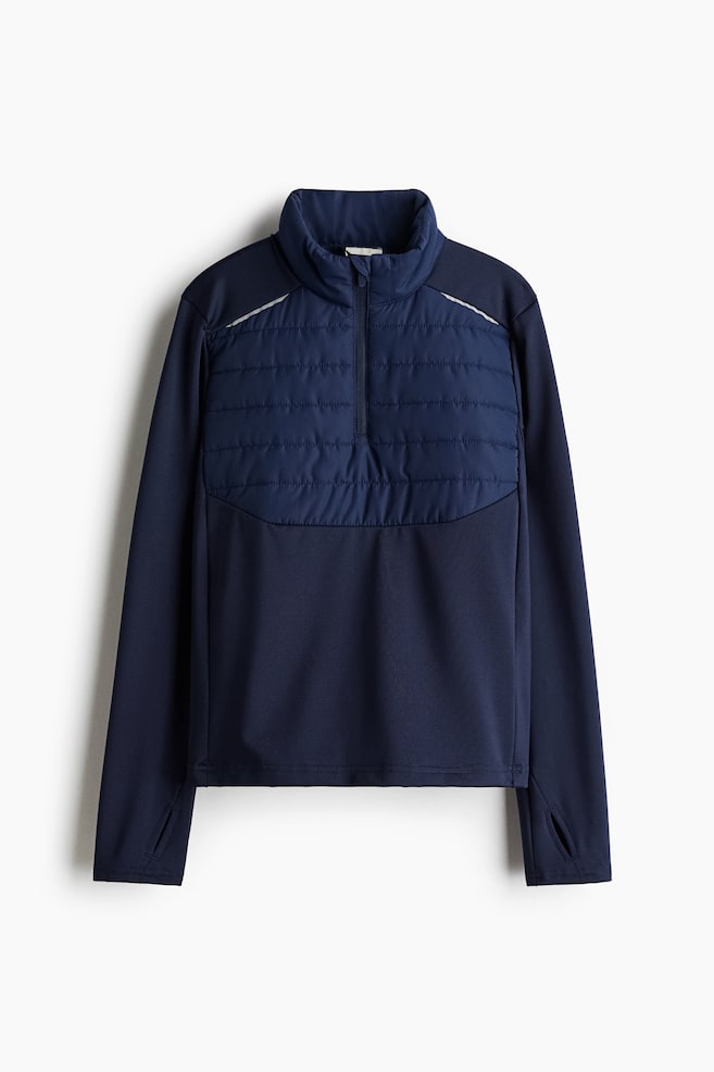 A dark blue football top is being presented from the front, featuring a high stand-up collar and a quarter-zip closure. It is designed with a quilted panel across the upper chest and shoulders, accented by thin reflective strips, and long sleeves featuring integrated thumbholes at the cuffs. - 1
