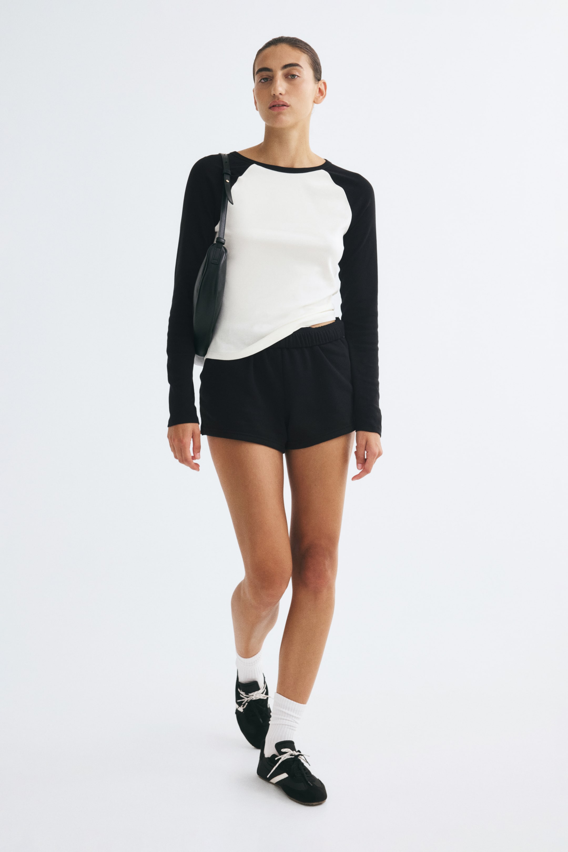 Visa större bild: En figursydd topp med vit kropp, rund halsringning och långa svarta raglanärmar bärs av modellen. Toppen är stylad med svarta shorts, en svart axelväska och svarta sneakers med vita strumpor.