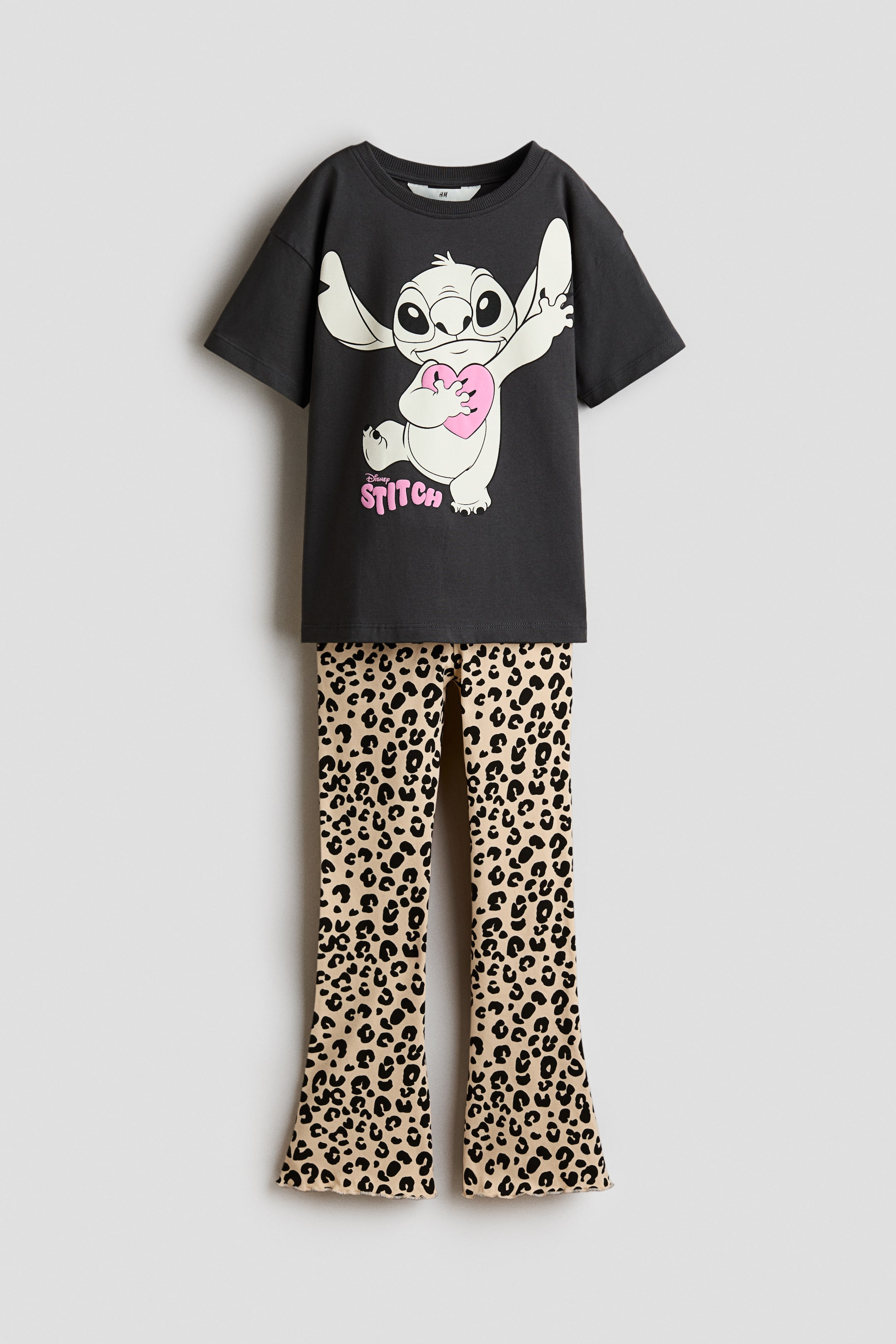 Visualizza immagine più grande: Un completo due pezzi composto da una t-shirt grigio scuro e pantaloni a gamba svasata con stampa leopardata beige e nera. La t-shirt presenta una grande grafica di Stitch che tiene in mano un cuore rosa con la scritta "Stitch" in rosa sotto, mentre i pantaloni sfoggiano un orlo arricciato.