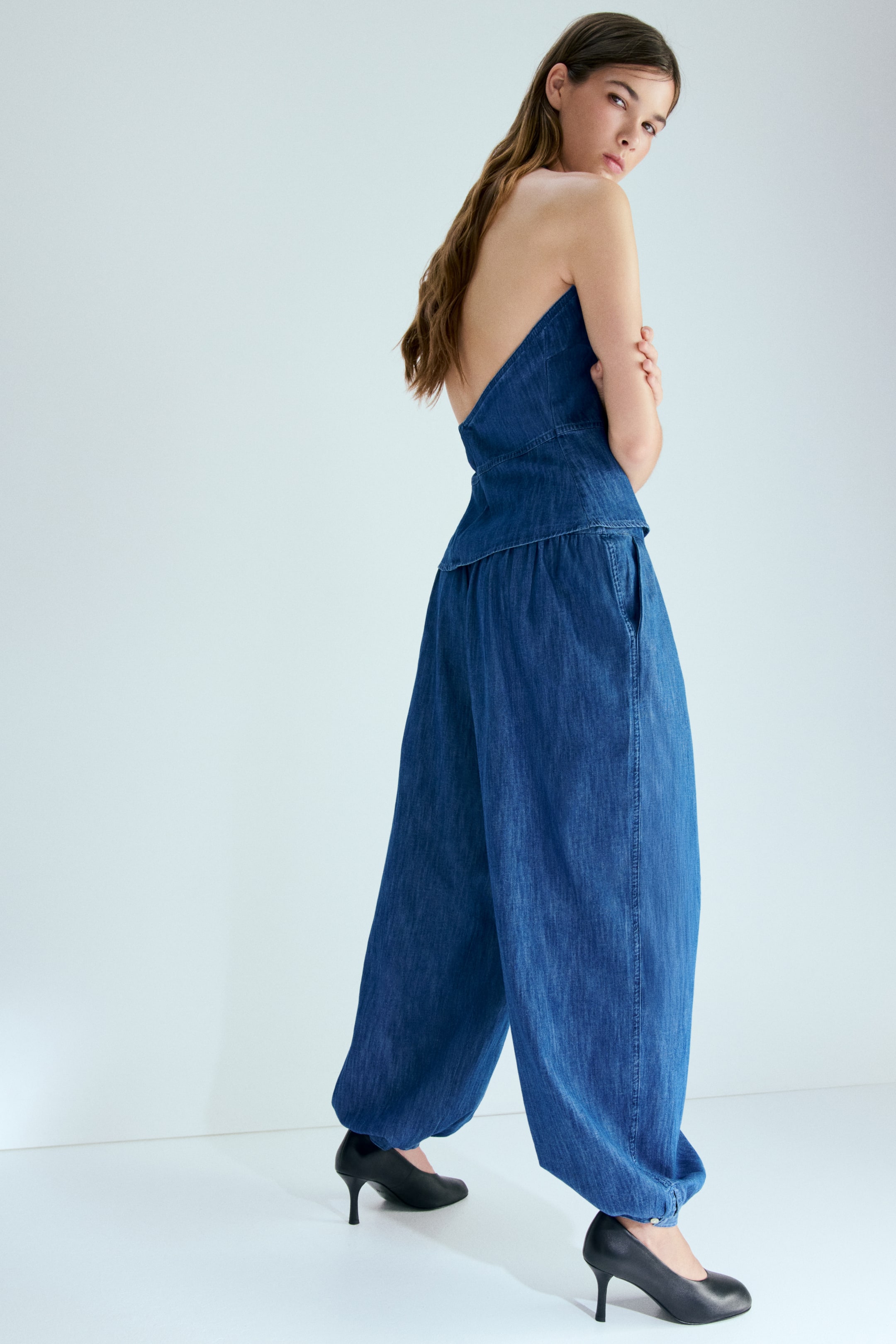 View larger image: A denim strapless top and high-waisted, wide-leg pants form a chic two-piece ensemble, showcased from a rear-left angle. The top boasts a diagonal backline and a fitted waist, while the pants highlight a gathered waist, side pockets, and a balloon silhouette with gathered cuffs resting elegantly above black high heels.