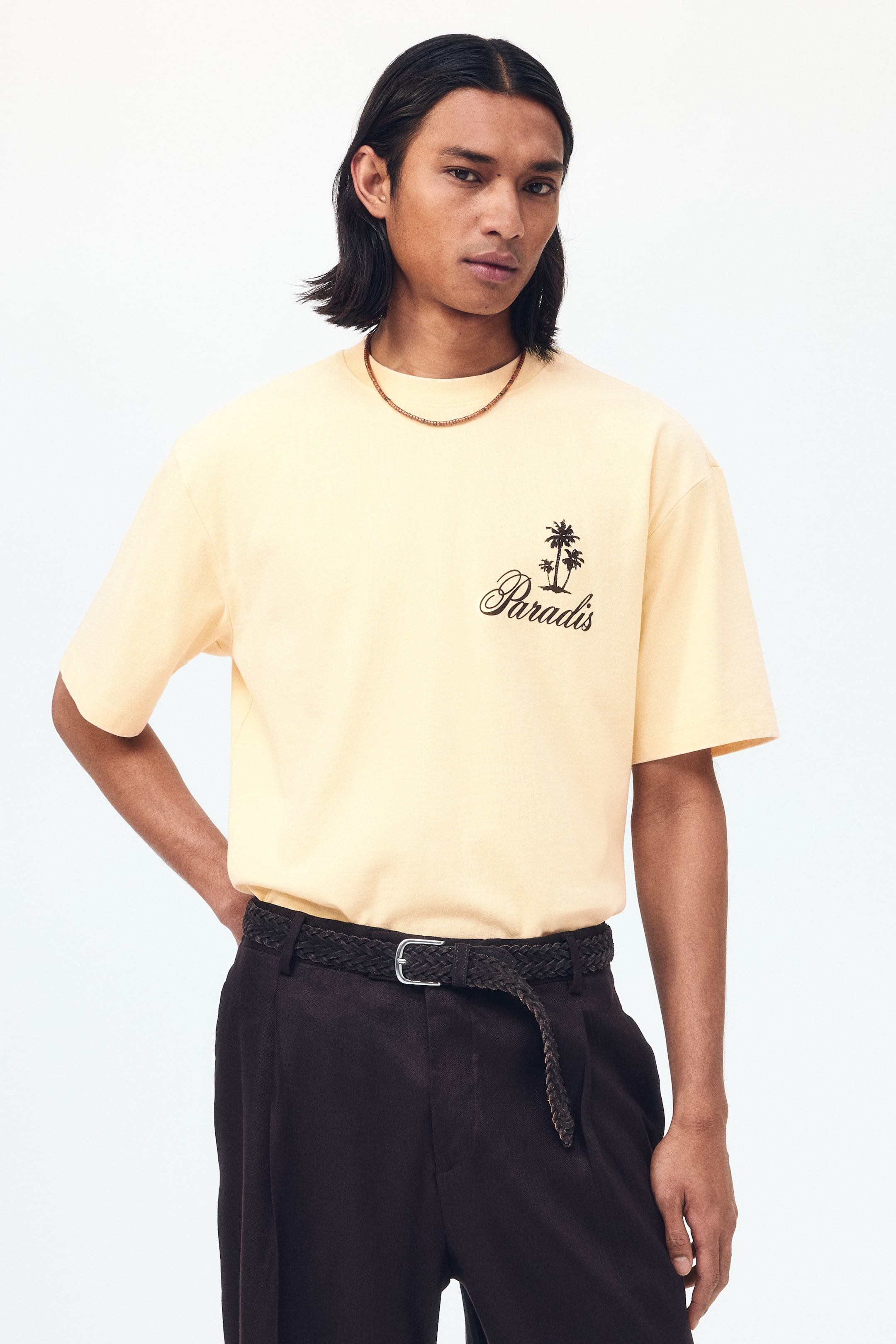 拡大画像を表示: ライトイエローのTシャツが正面向きに紹介されています。リラックスフィットとショートスリーブが特徴です。左胸には黒いグラフィックがあり、スクリプトフォントで描かれた「Paradis」という言葉と、2本のヤシの木のシルエットが描かれています。