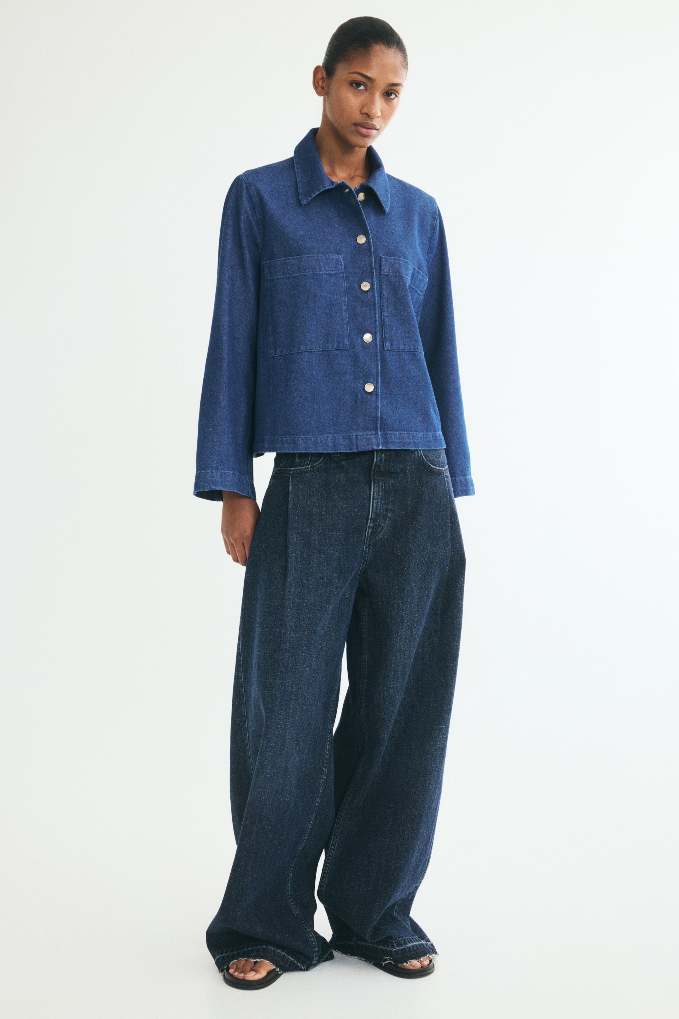 View larger image: A short blue denim jacket, shown from the front, features a classic collar, metal button closures down the front, and two large patch chest pockets. The jacket has wide sleeves and a straight-cut hem, styled with dark blue wide-leg jeans.