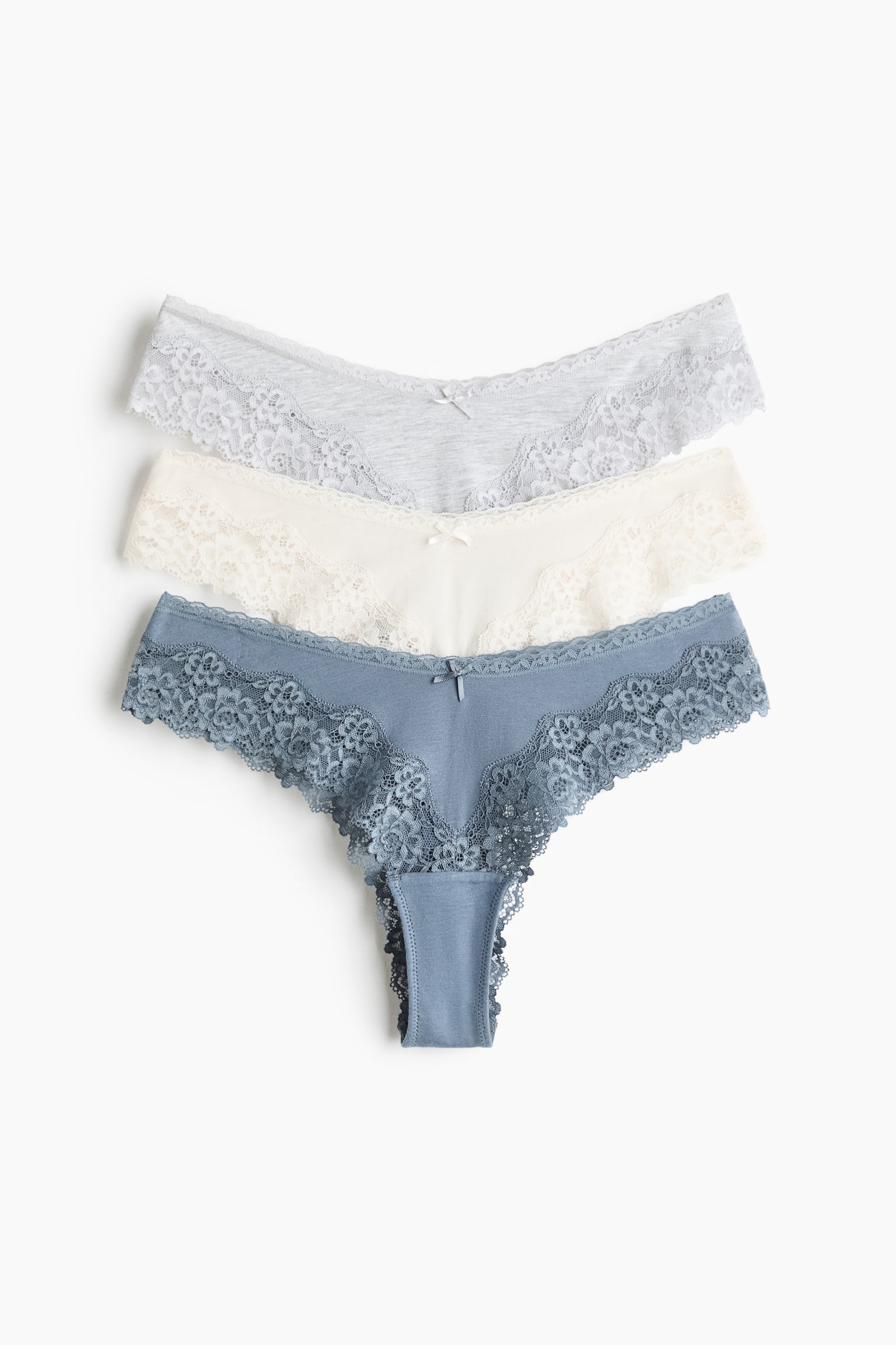 Trois paires de slips taille basse en gris clair, crème et bleu poudré sont disposées à plat en pile, montrant la vue de face. Chaque paire présente un corps en jersey de coton avec une taille en V, un petit nÅud décoratif au centre et de larges empiècements en dentelle festonnée sur les côtés.