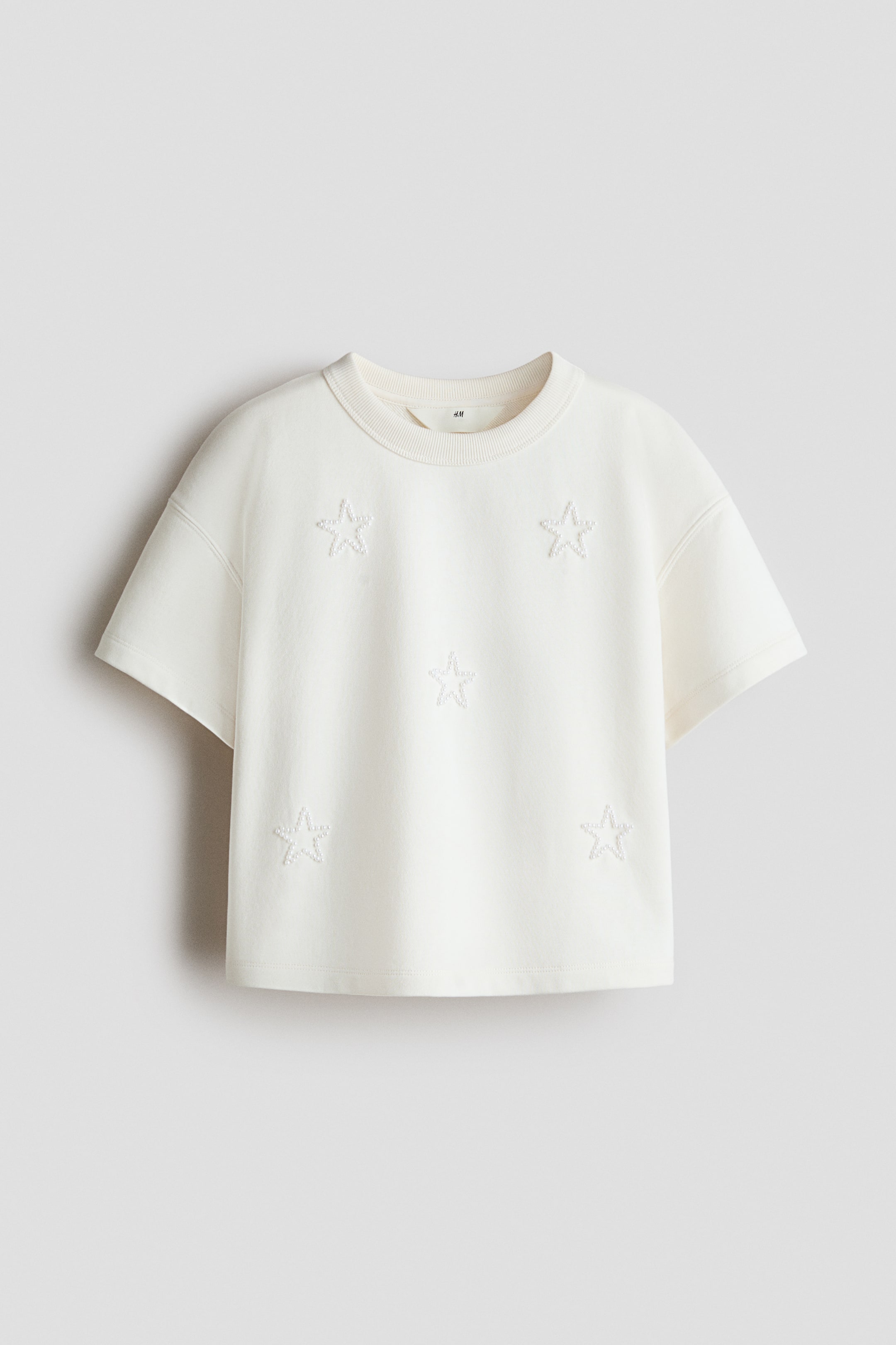 Größeres Bild anzeigen: Ein cremeweißes, kurzärmeliges Jersey-T-Shirt wird von vorne präsentiert, mit einem runden, gerippten Halsausschnitt, überschnittenen Schultern und gestickten Sternumrissen, die die Vorderseite zieren.