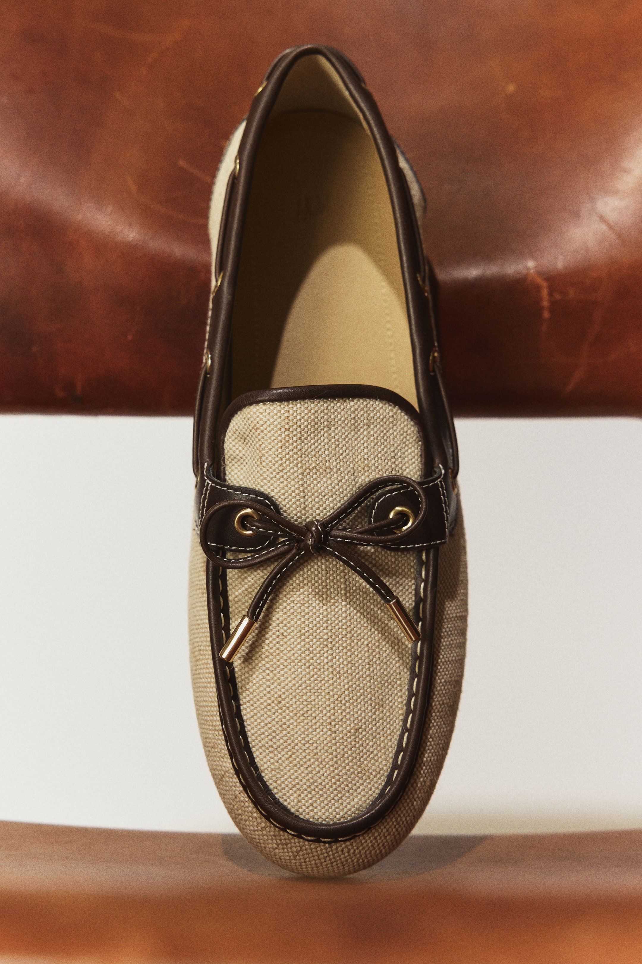 Größeres Bild ansehen: Ein Loafer steht aufrecht, aus leicht erhöhter Draufsicht betrachtet, und verfügt über ein beigefarbenes, gewebtes Obermaterial mit dunkelbraunen Lederbesätzen. Dunkelbraune Lederbänder bilden auf dem Spann (Vamp) eine dekorative Schleife, akzentuiert durch weiße Kontrastnähte und goldfarbene Aglets.