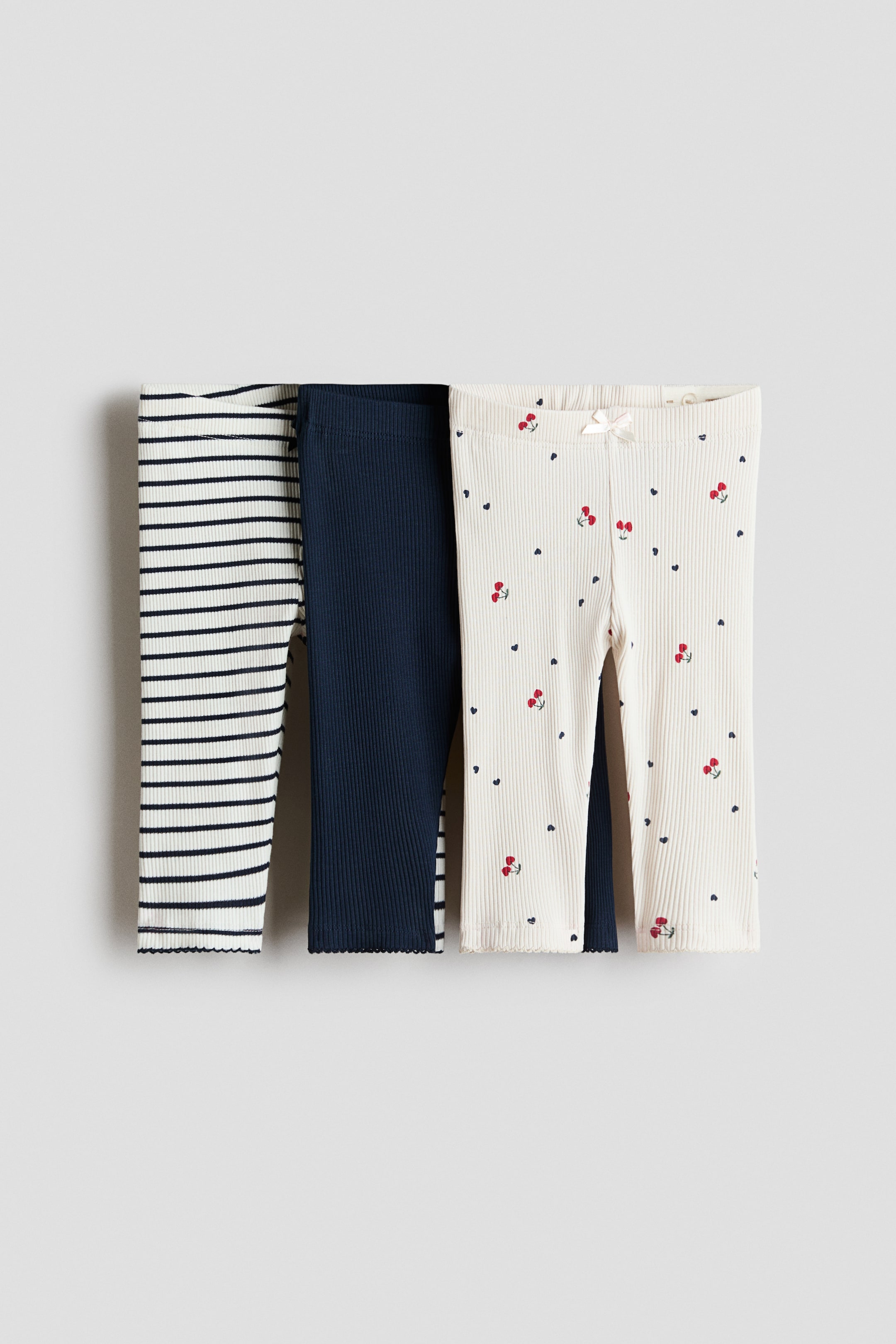 Ver imagen más grande: Tres pares de leggings de bebé acanalados se presentan desde el frente, cada uno con aberturas en las piernas con borde festoneado. Un par muestra rayas horizontales en azul marino sobre un fondo crema, otro es de color azul marino sólido, y el tercero presenta un fondo crema con pequeños corazones en azul marino y cerezas rojas, acentuado por un pequeño lazo crema en la cintura.