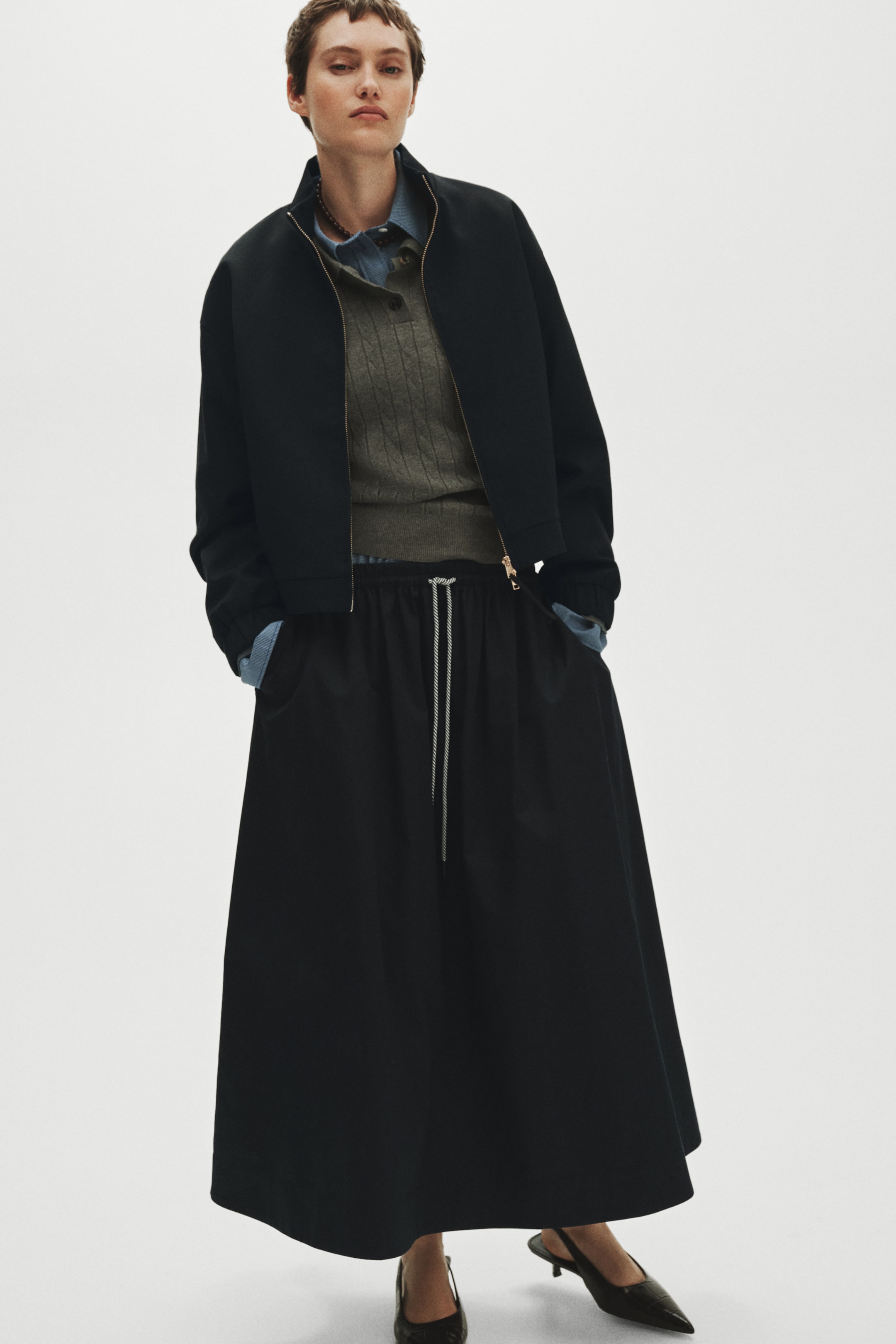 Grösseres Bild ansehen: Eine schwarze Bomberjacke zeichnet sich durch einen durchgehenden goldfarbenen Reissverschluss aus und wird über einem olivgrünen, gerippten Pullover sowie einem blauen Denimhemd mit hochgestelltem Kragen getragen. Ein fliessender schwarzer Maxirock weist einen sichtbaren weiss-schwarzen Kordelzugbund und Seitentaschen auf und wird mit schwarzen, spitzen Slingback-Heels kombiniert.