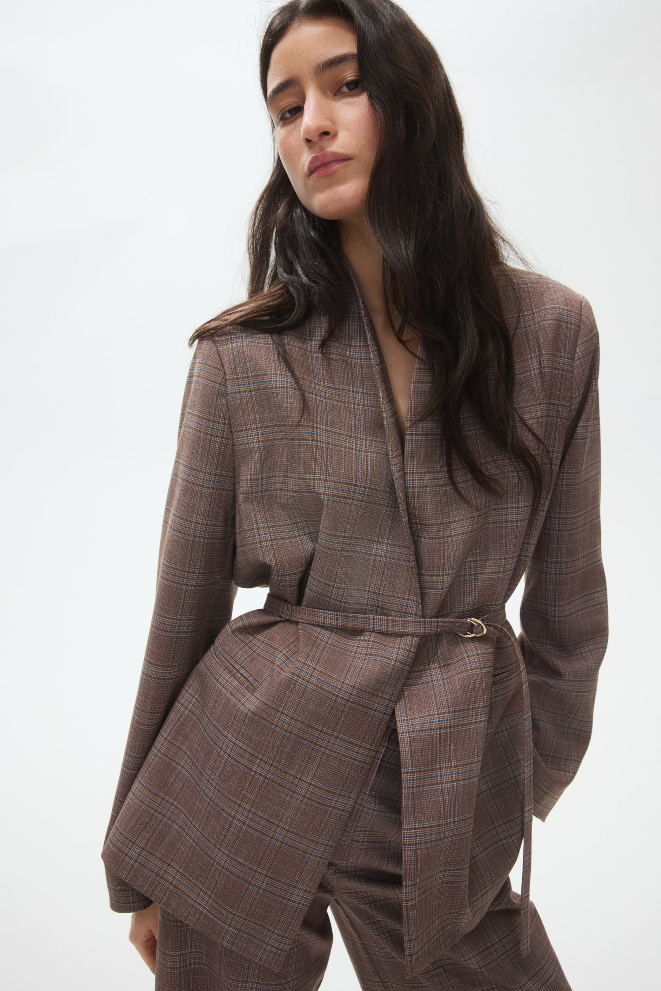 View larger image: The model is wearing a brown plaid blazer crafted from woven fabric, showcasing a lapelless V-neckline, structured shoulders, and a subtly overlapping front. A slim, detachable belt with D-rings cinches the waist.