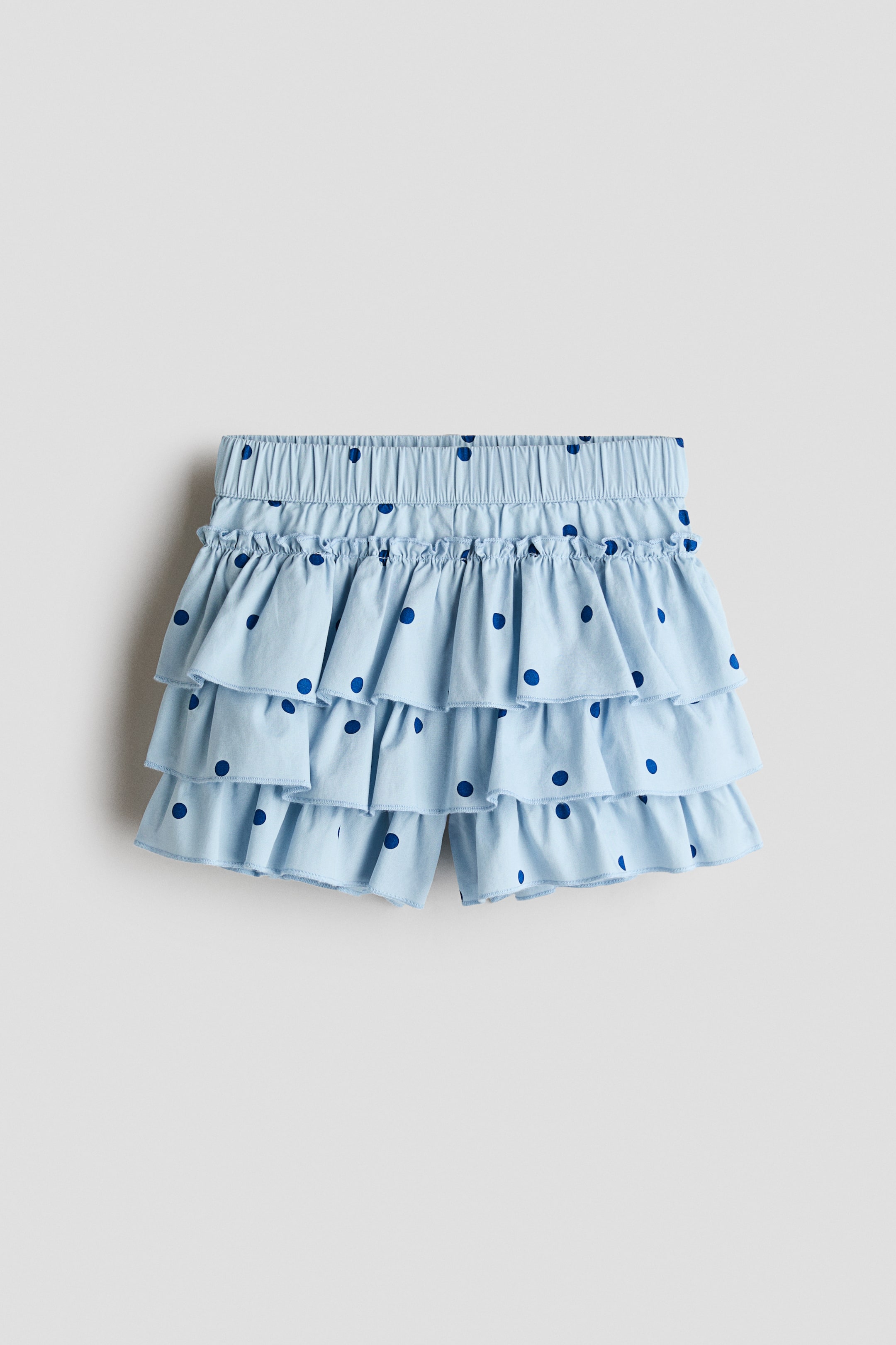 View larger image: A light blue tiered skort with dark blue polka dots is shown from the front, highlighting an elasticated waistband with a frill trim. The design features three tiers of ruffles draping over the integrated shorts, which are visible at the bottom hem.