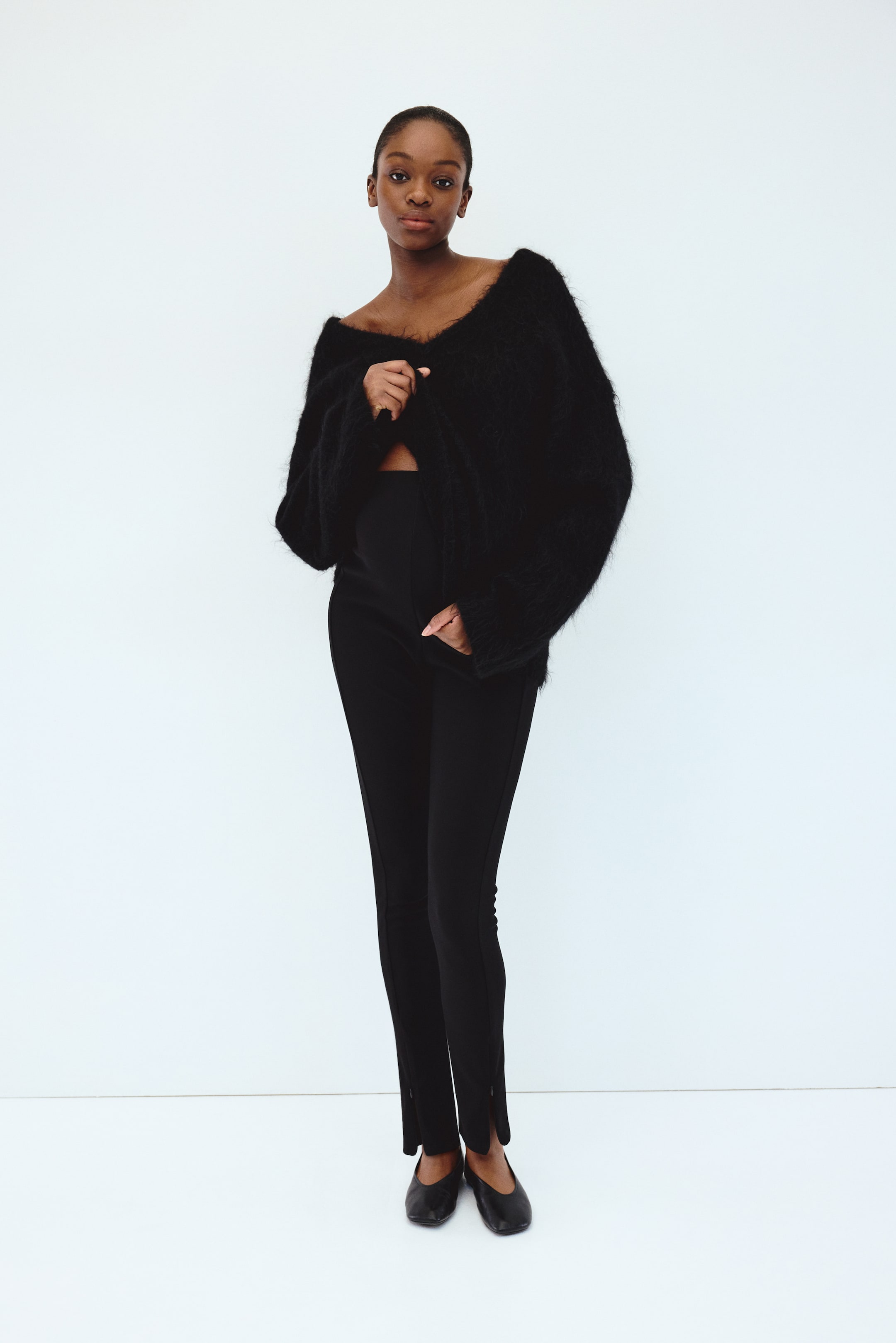 Grotere afbeelding bekijken: Een zwarte, pluizige cardigan valt van de schouders en heeft een brede V-hals en lange, losse mouwen. Hij wordt gedragen over een zwarte, hooggetailleerde slim-fit broek met gesplitste zomen bij de enkels, met zwarte ballet flats aan de voeten.
