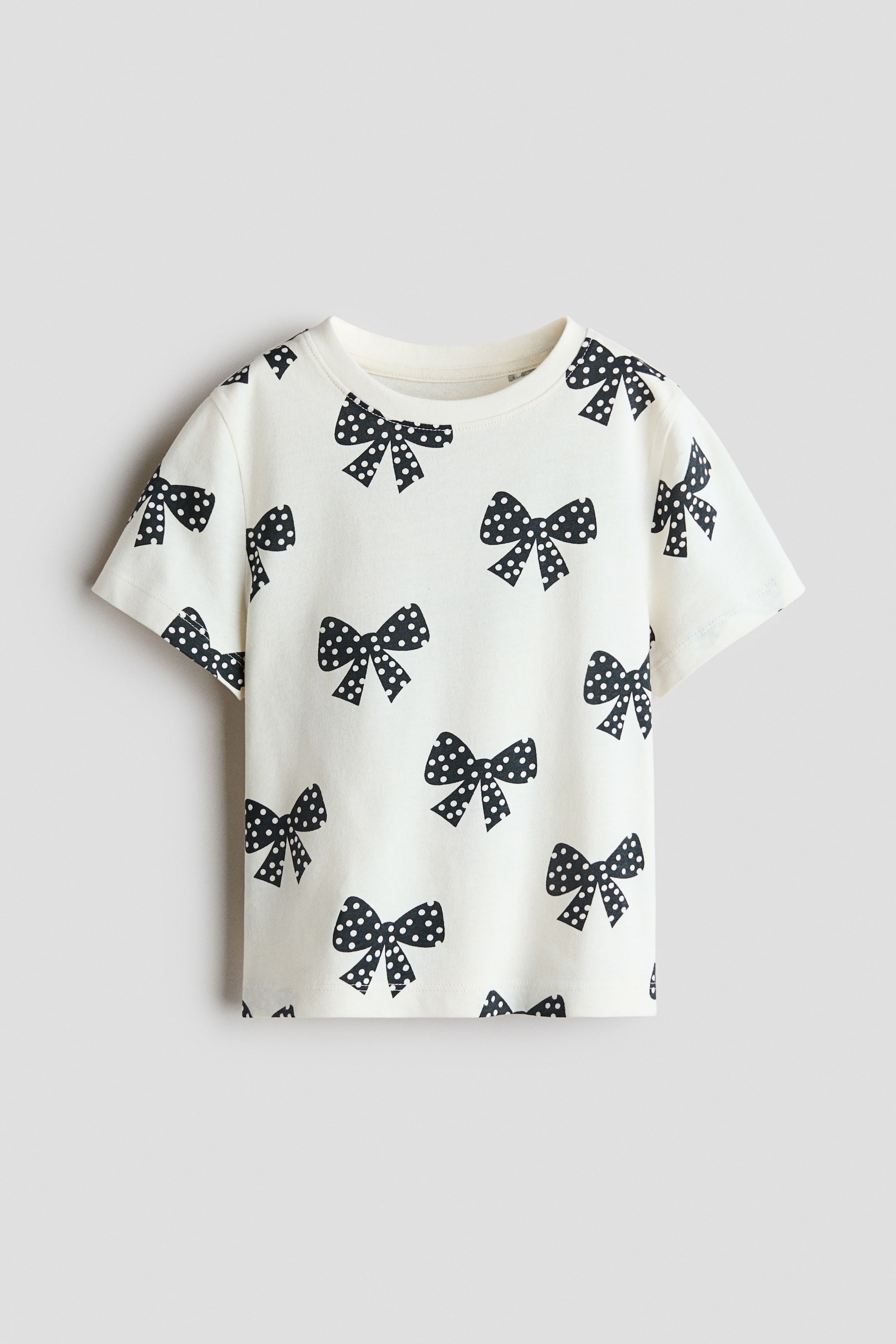 View larger image: An off-white cotton jersey T-shirt is showcased from the front, featuring an all-over print of black bows with white polka dots. The tee features a round, rib-trimmed neckline, gently dropped shoulders, and short sleeves.
